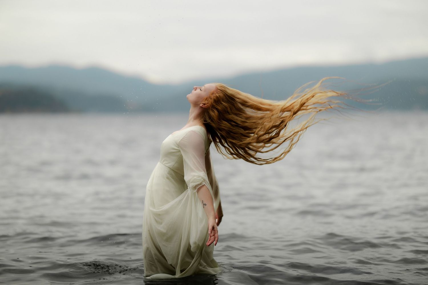 A woman in a white dress stands in a lake, her long red hair flowing dramatically in the wind against misty mountains.