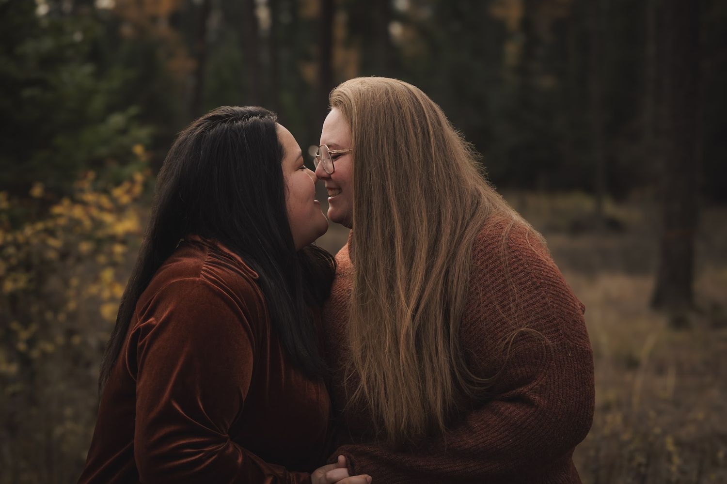 Two women share a romantic moment in an autumn forest, leaning in for a kiss wearing warm earth-toned sweaters.