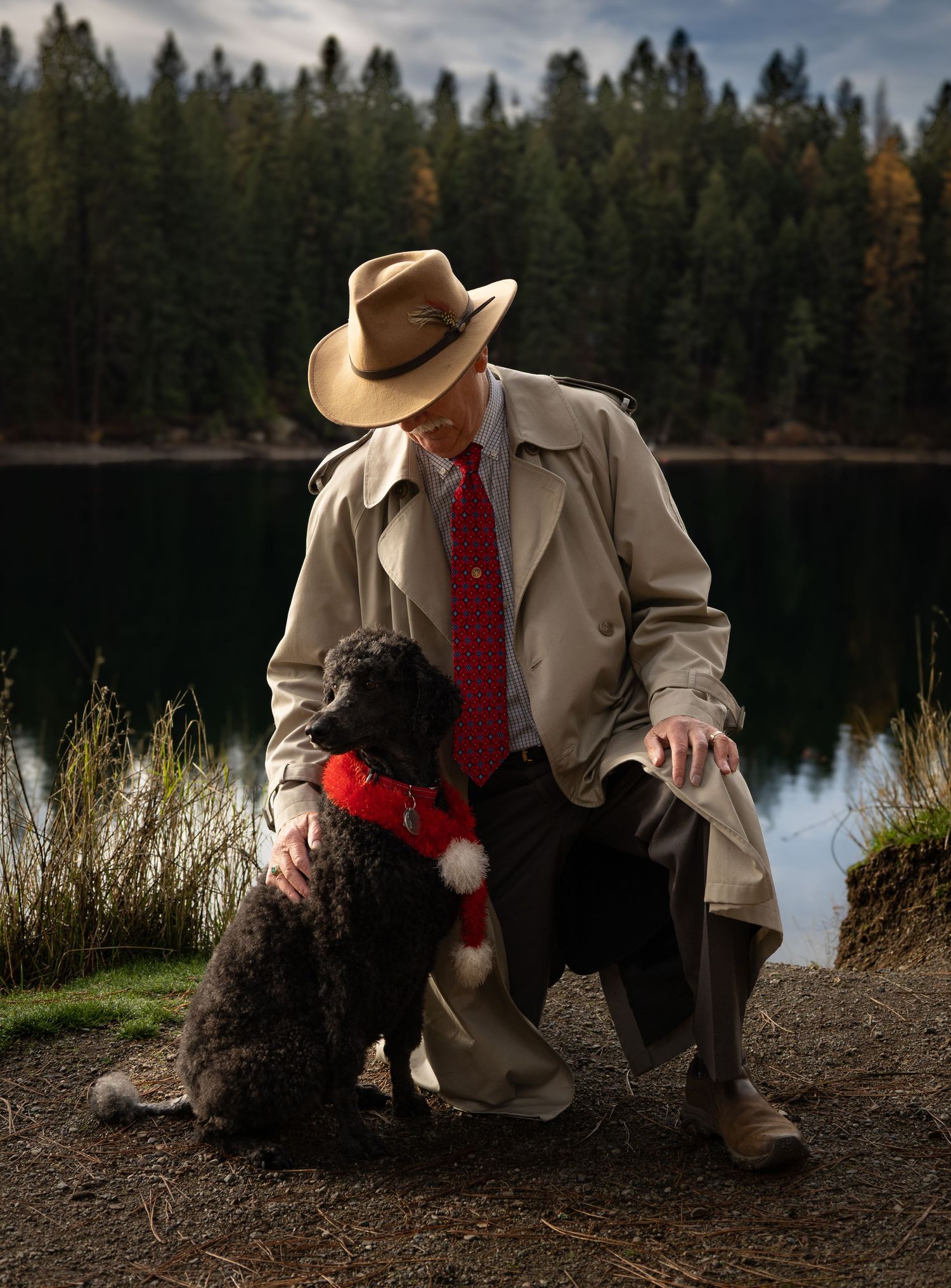 Elderly man in tan hat and red tie kneeling beside a curly black dog near a reflective forest lake.