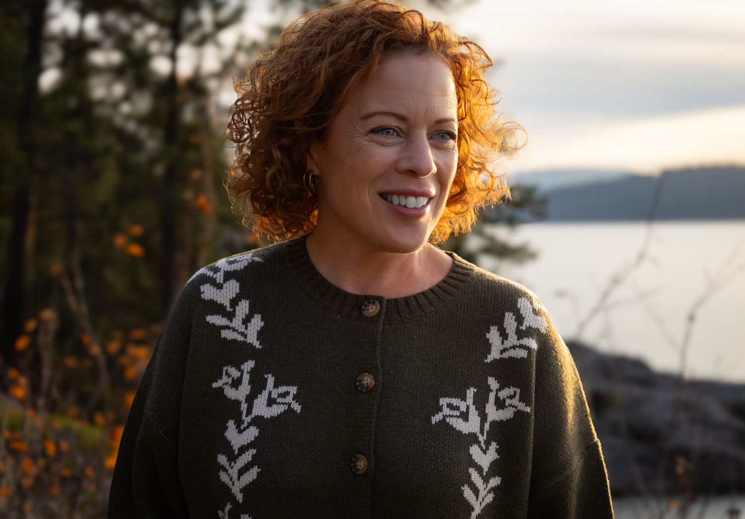 Woman with curly red hair smiling outdoors, wearing a dark floral cardigan sweater near a lake at sunset.
