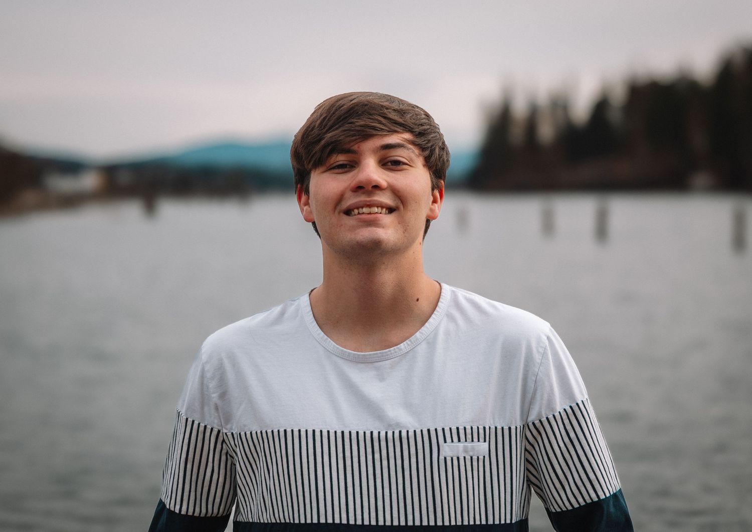 Smiling teenage boy in striped t-shirt standing outdoors near a lake with mountains and trees in background.