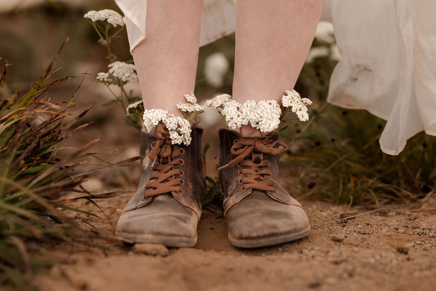 Rustic brown lace-up boots adorned with delicate white wildflowers on a dirt path, paired with a white dress.