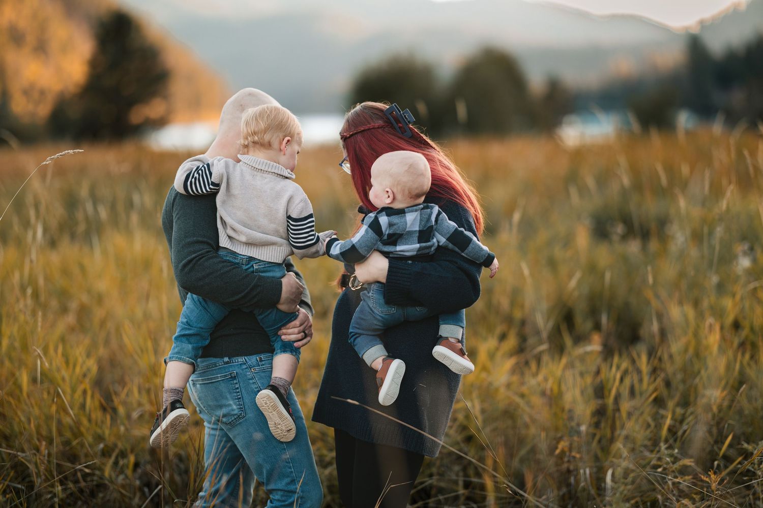 Two parents holding toddlers in a golden field with mountains, sharing a tender family moment at sunset.