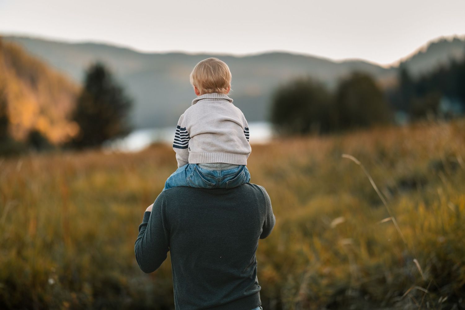 A father carries his toddler on his shoulders in a scenic autumn field with mountains and a lake in the background.