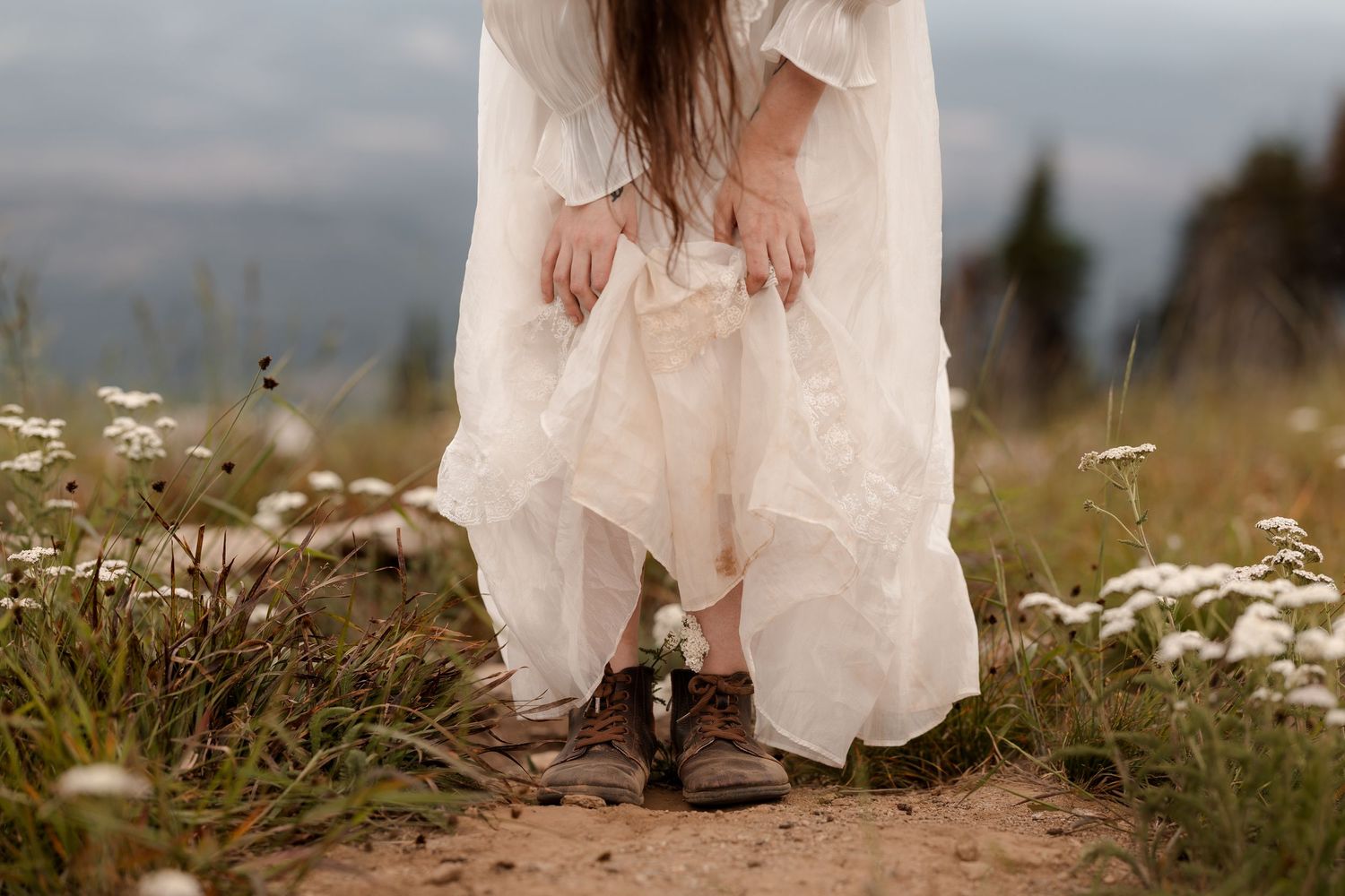Bride in white lace dress wearing rustic brown boots, bending down among wildflowers in a meadow.