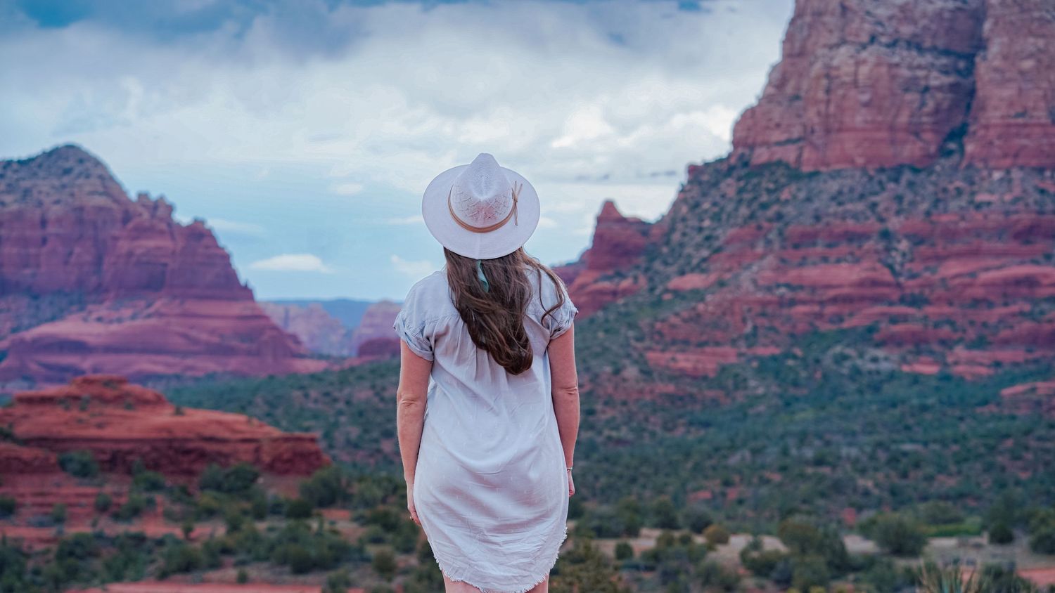 Woman in white dress and hat gazes at stunning red rock formations in Sedona, Arizona landscape.