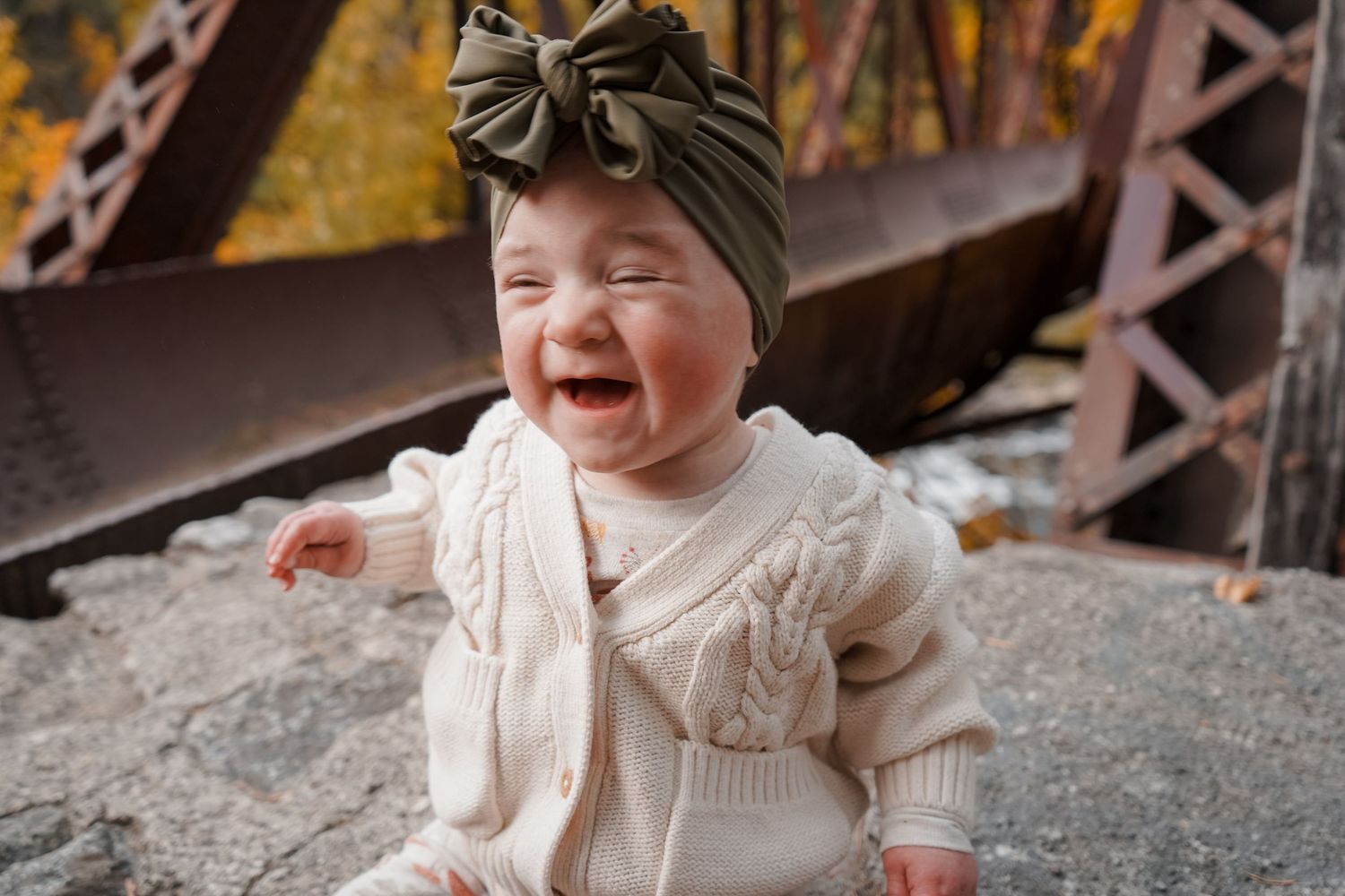 Laughing baby in cream knit sweater and olive bow turban headband, sitting outdoors near rustic bridge in autumn.
