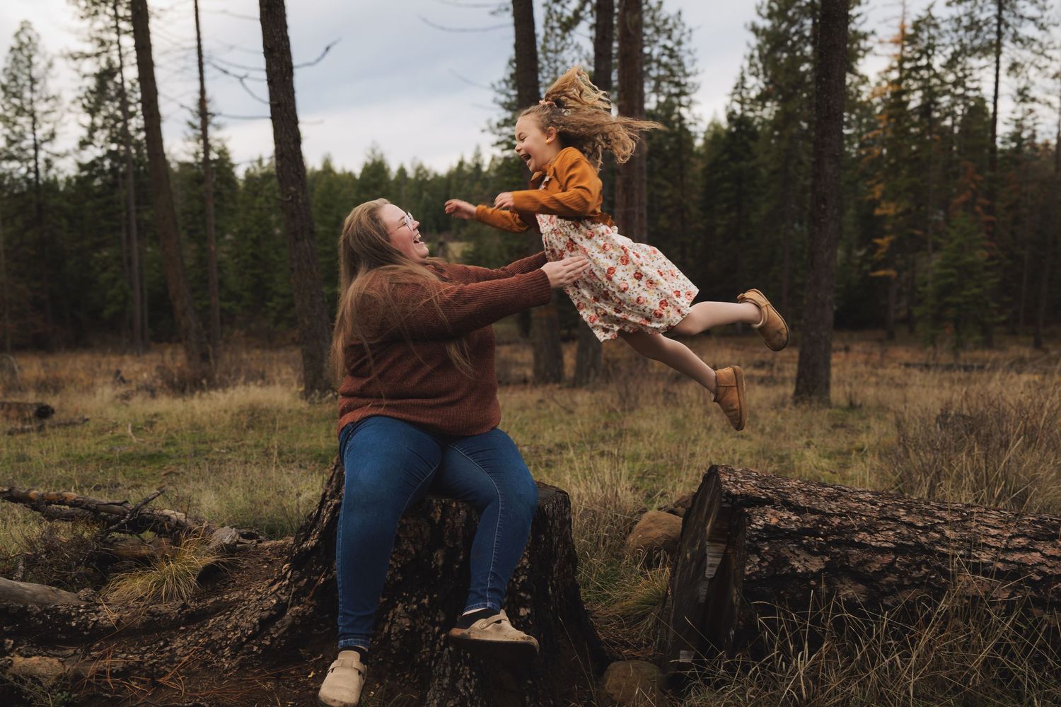 A mother sits on a log in a forest, playfully tossing her young daughter into the air.