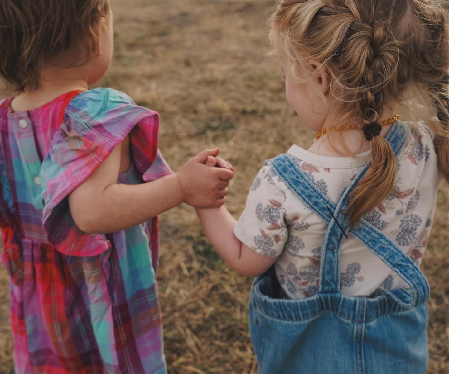 Two young girls holding hands in a field, one wearing a tie-dye dress and the other in denim overalls.