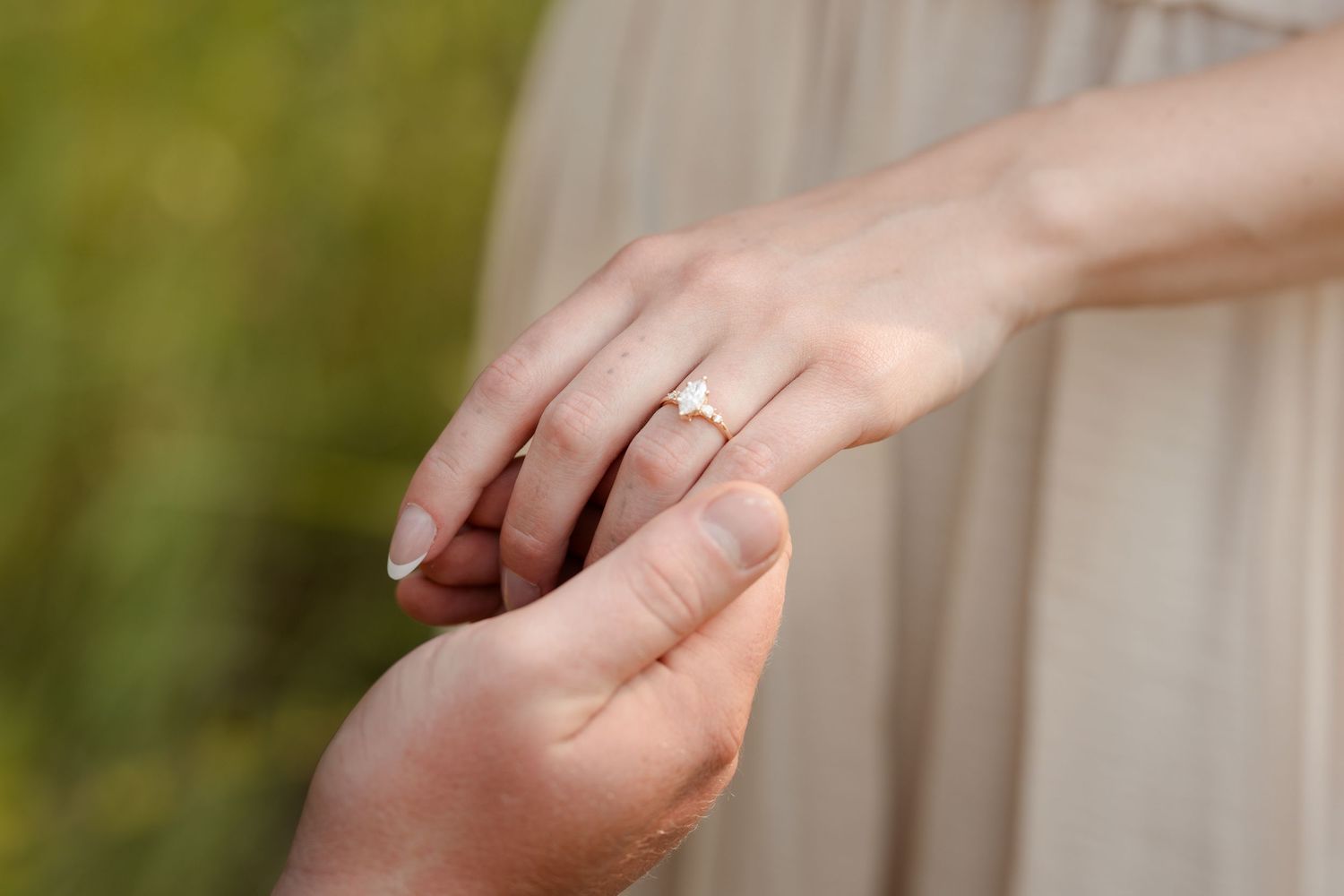 Close-up of a woman's hand wearing a diamond engagement ring while holding hands with a partner outdoors.