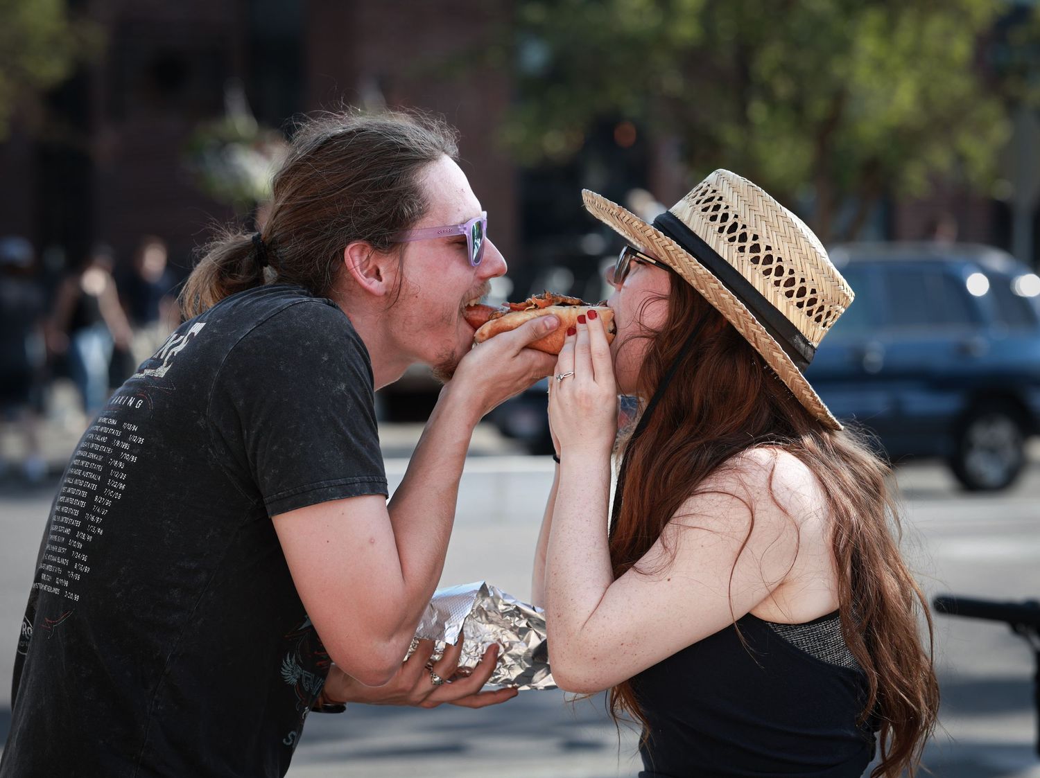 Couple sharing a slice of pizza outdoors on a sunny day, woman wearing straw hat and sunglasses.