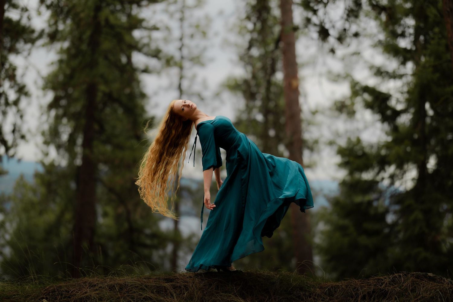 A red-haired woman in a flowing teal gown dances gracefully among tall forest trees.