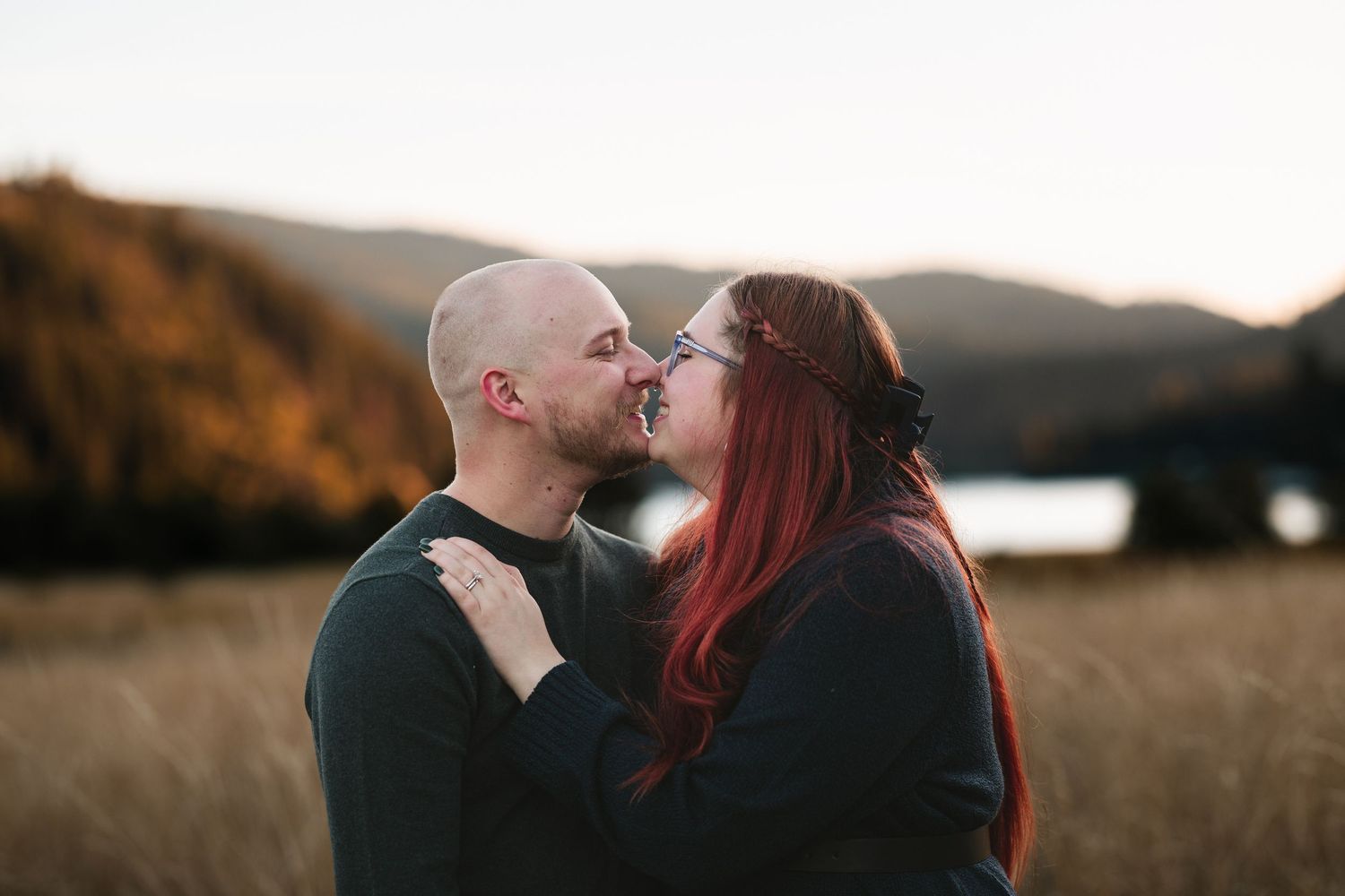 Couple sharing a romantic kiss in a golden field with mountain and lake backdrop during sunset.