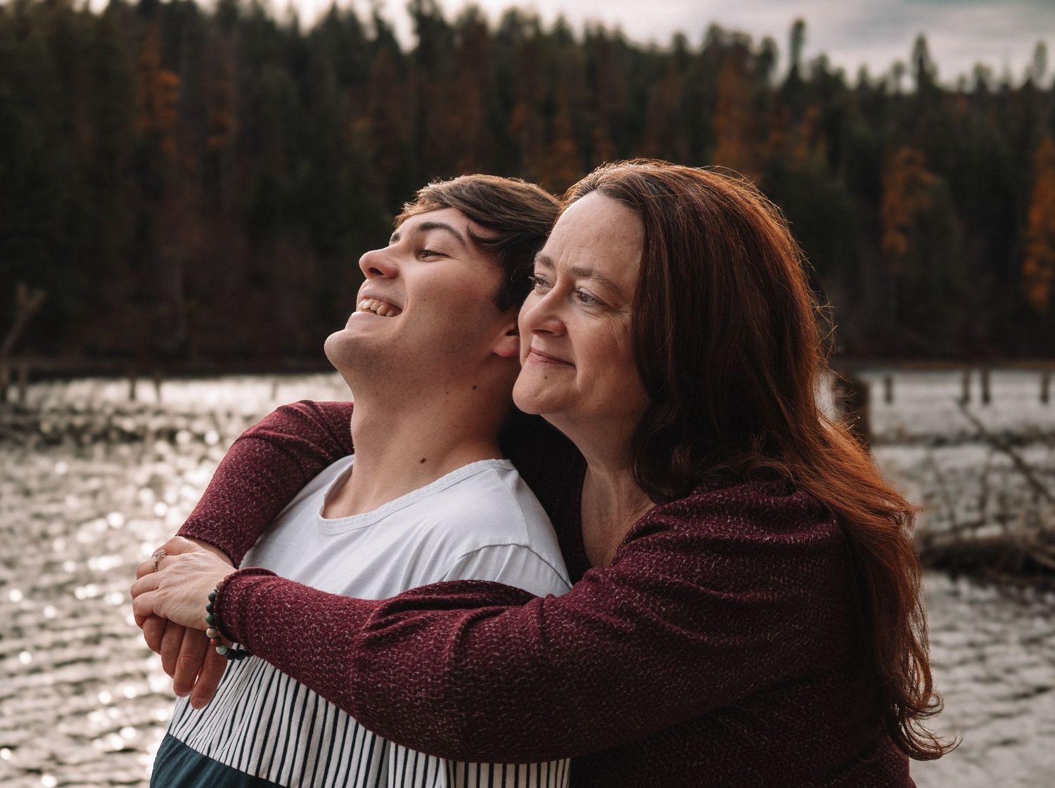 Mother and son hugging and smiling by a lake surrounded by autumn trees in a warm outdoor portrait.