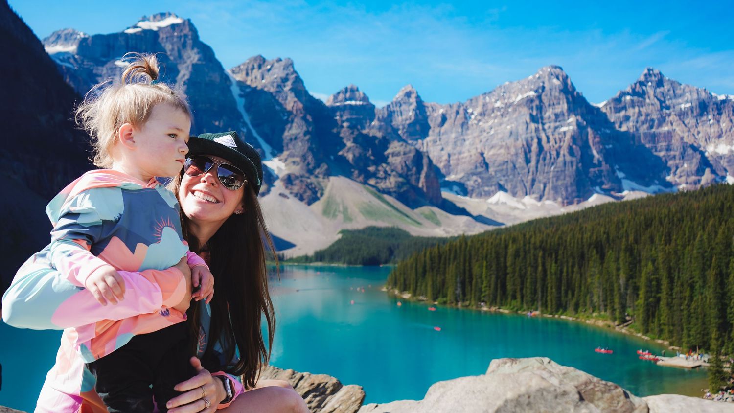 A mother holds her toddler while enjoying the stunning turquoise lake and mountain views.