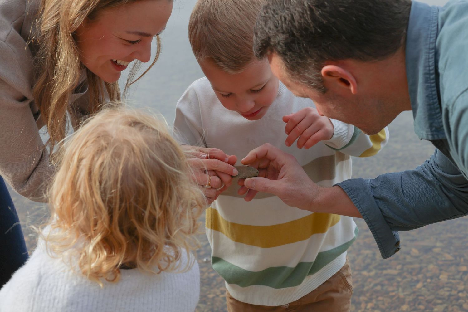 Family of four examining a small shell together outdoors, smiling and bonding near a beach.