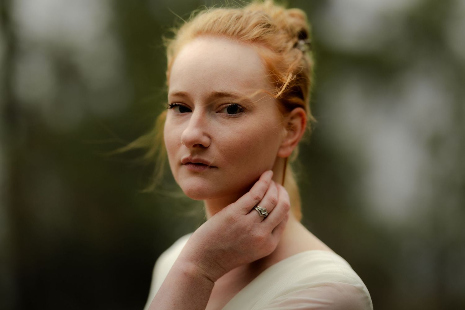 Young woman with red hair in updo, wearing a ring, posing outdoors with a blurred green background.
