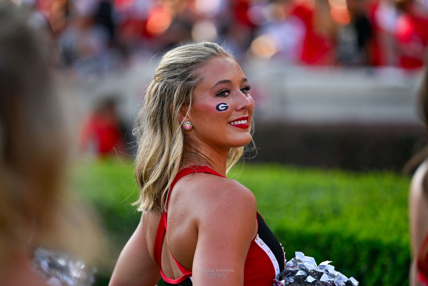 Smiling cheerleader with face paint at a college football game, wearing a red and black uniform with pom-poms.