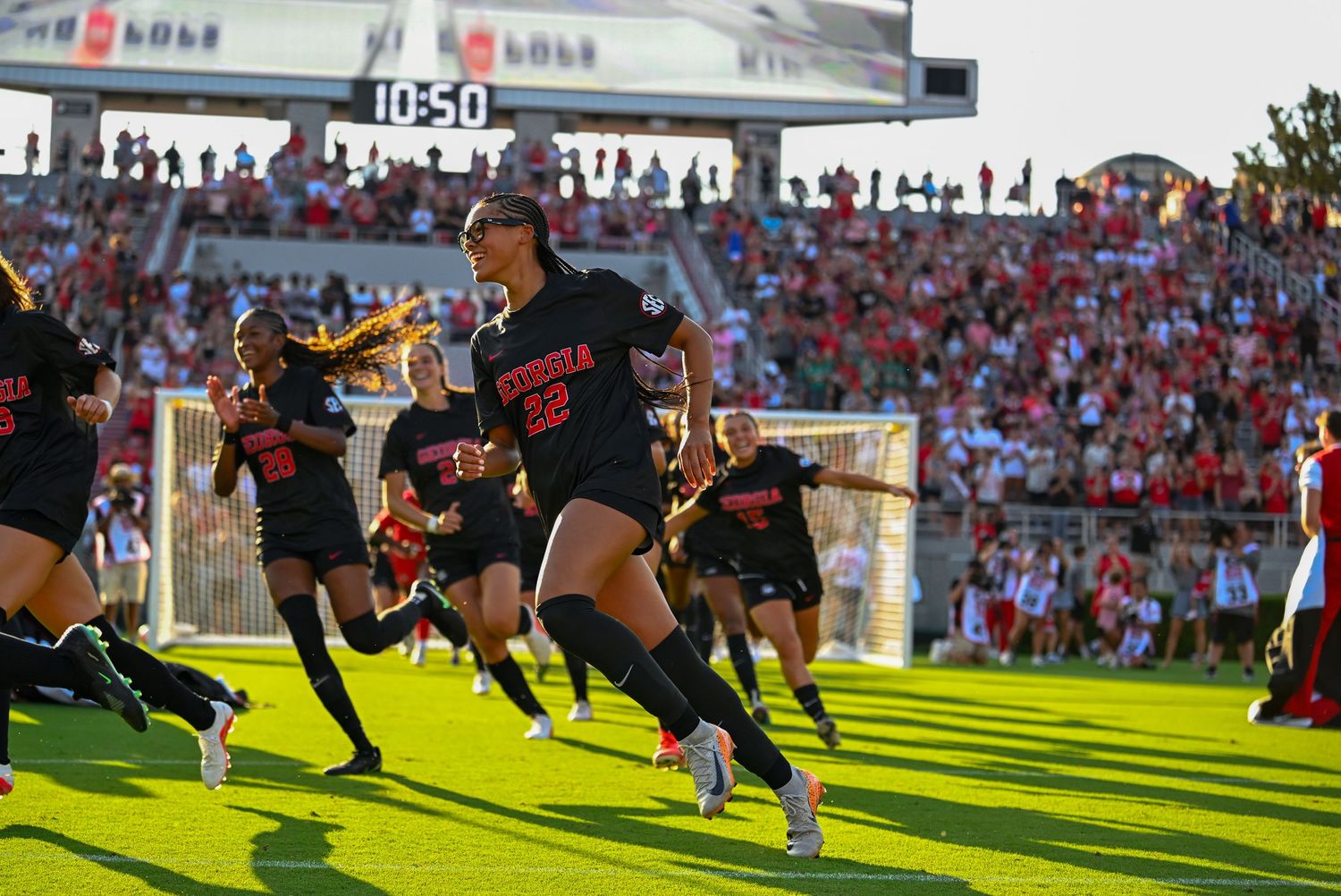 Female soccer players in red uniforms sprint onto a sunlit field before a packed stadium crowd.