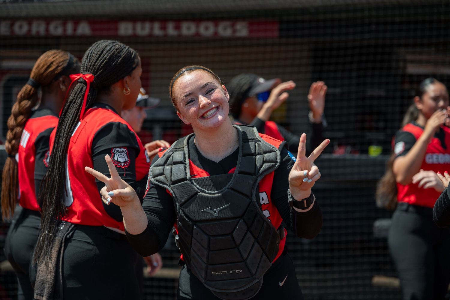 Georgia Bulldogs softball catcher in gear flashing peace signs, smiling at camera during game day.
