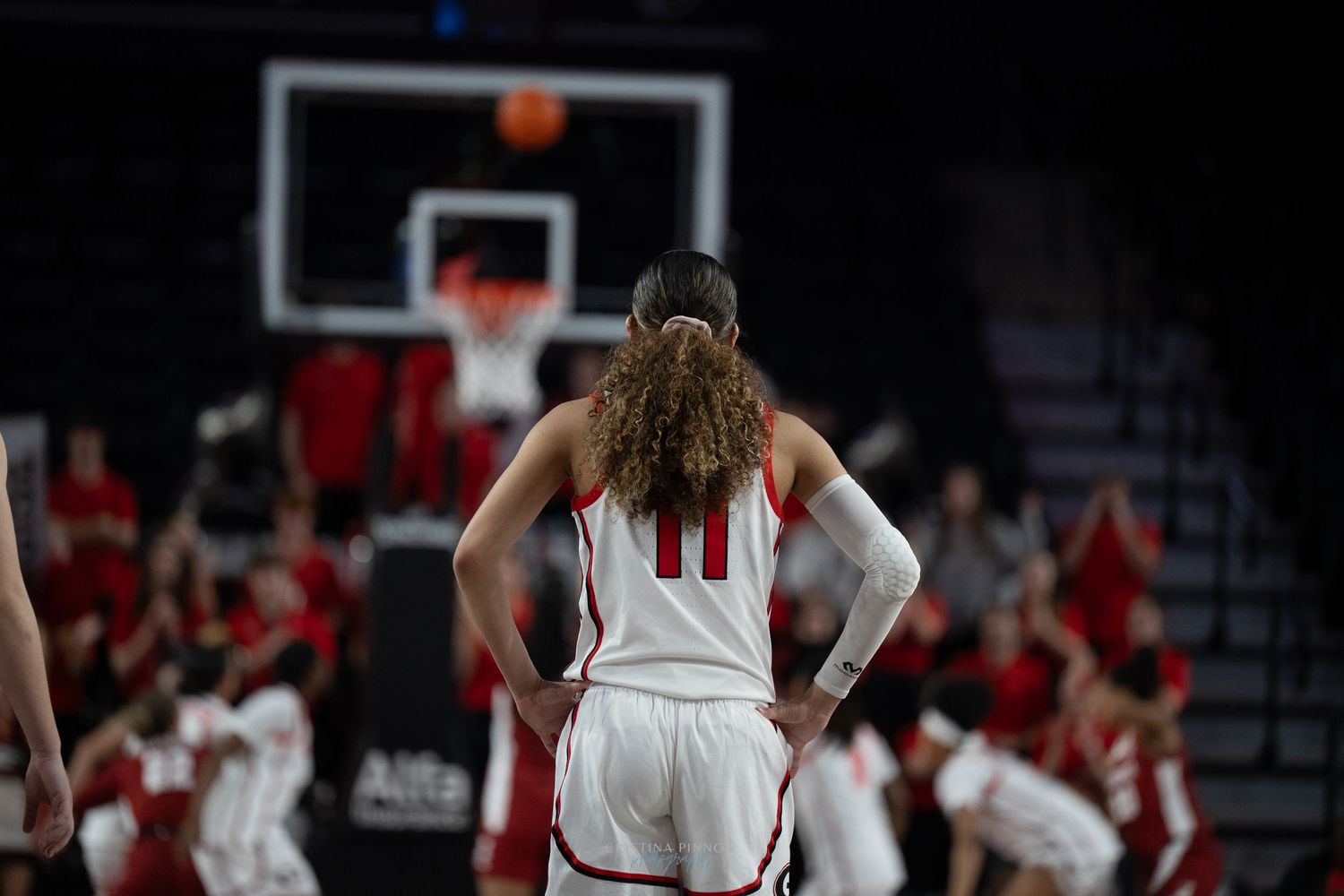 Basketball player in white uniform number 11 stands with hands on hips watching shot attempt during game.