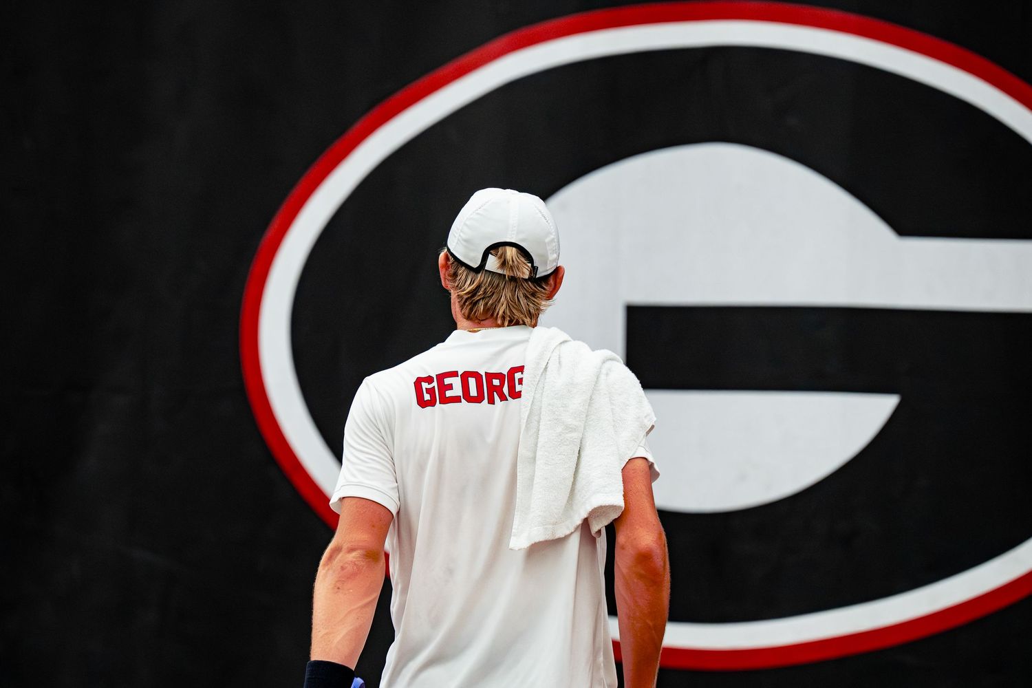 Georgia Bulldogs tennis player wearing white uniform and cap stands before large UGA logo on court.