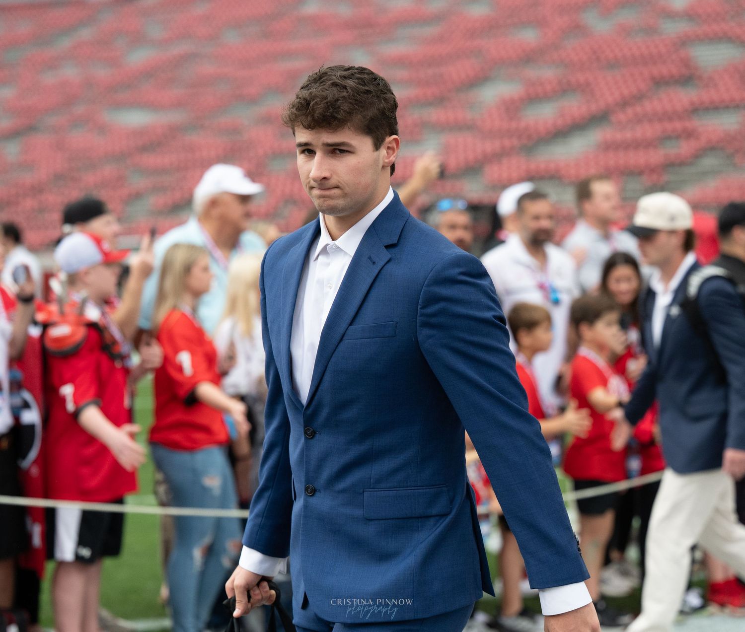 A person wearing a navy blue suit jacket stands in front of a stadium crowd.
