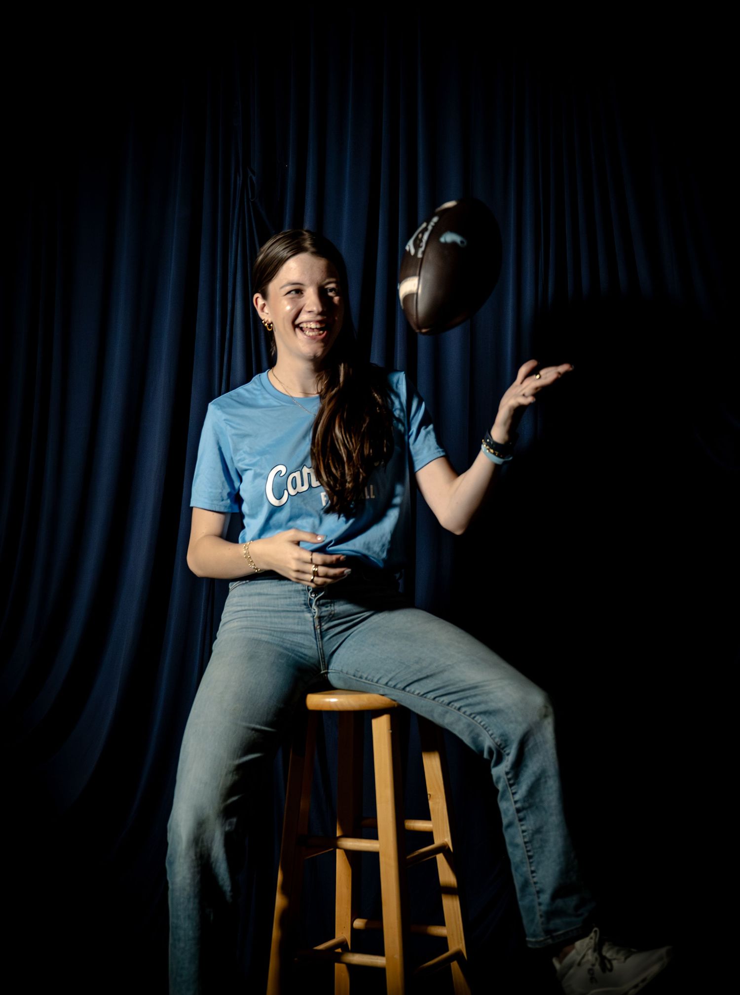 A smiling woman in a blue t-shirt tosses a hat while sitting on a wooden stool against a dark blue curtain.