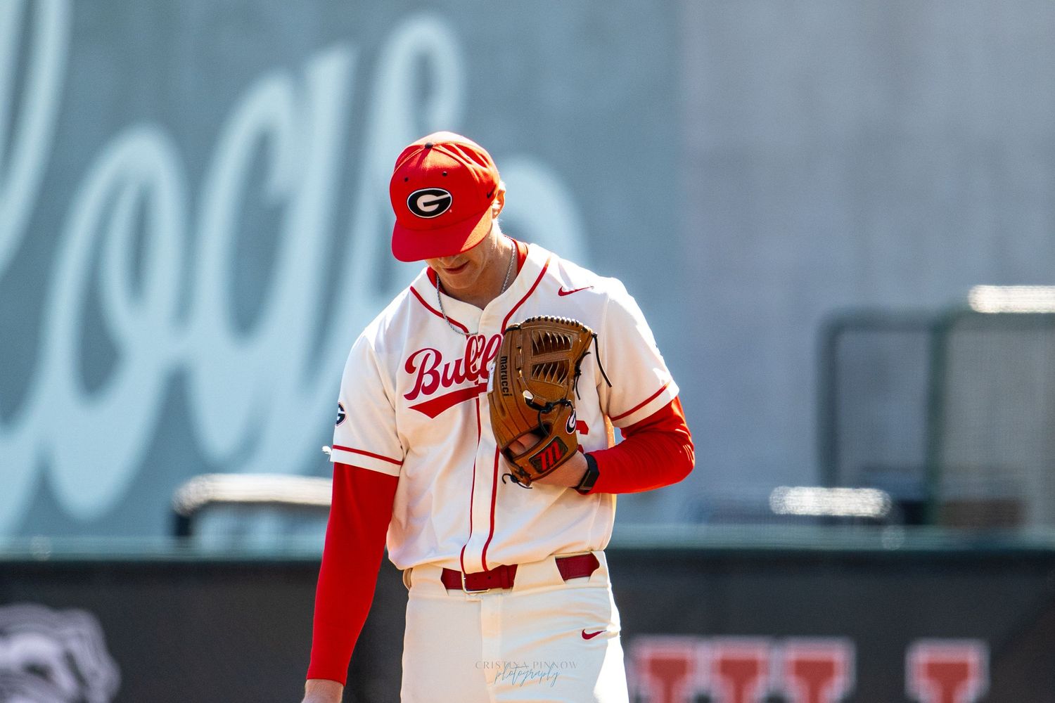 Georgia Bulldogs baseball player in white uniform and red helmet stands on field holding a glove during a game.