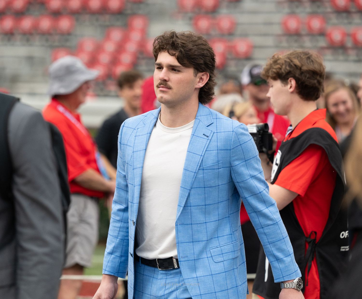 A person in a light blue blazer and white shirt stands in a stadium setting.
