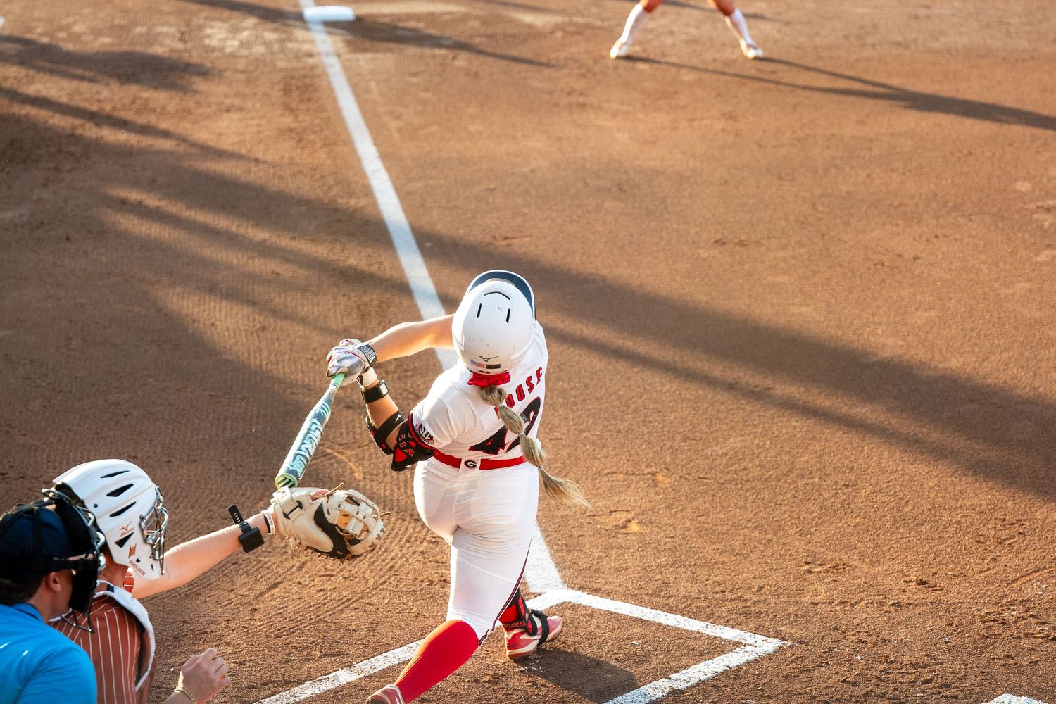 Softball batter in red uniform swings at pitch while catcher crouches behind home plate on dirt field.