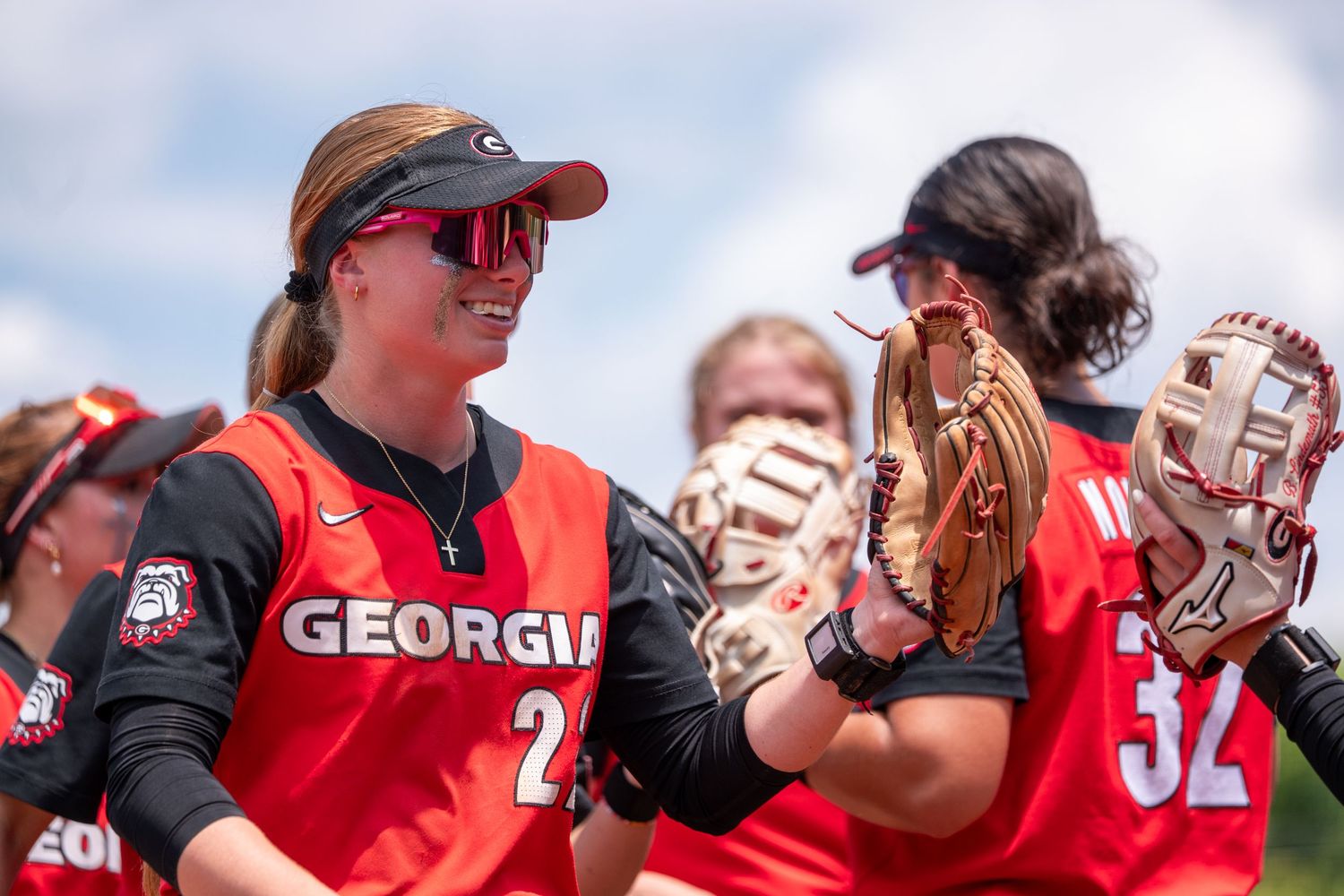 Georgia Bulldogs softball players in red uniforms celebrate with high-fives during a sunny outdoor game.