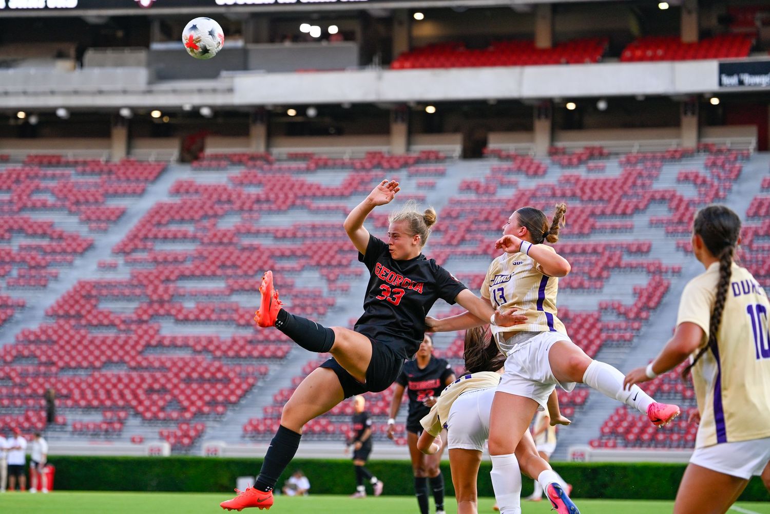 Female soccer player performs a high kick during a match in a stadium with purple and red team uniforms.