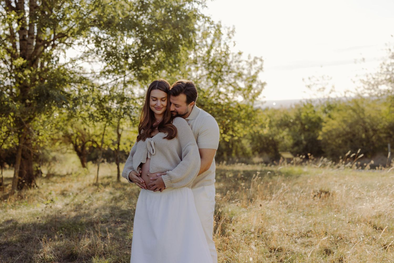 A couple embraces tenderly in a sunlit meadow, the woman in a white dress cradling her baby bump.