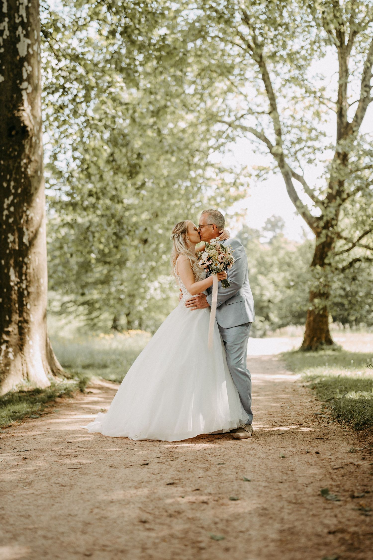 Bride and groom kissing on sun-dappled tree-lined path, wedding photography.