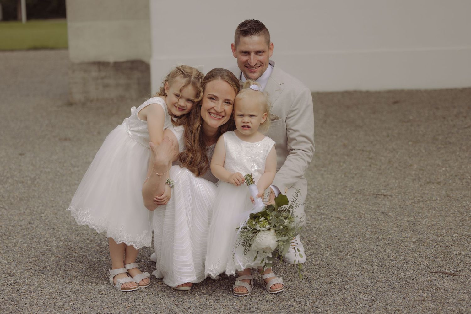 Smiling family with two little girls in white dresses posing outdoors at a wedding venue.
