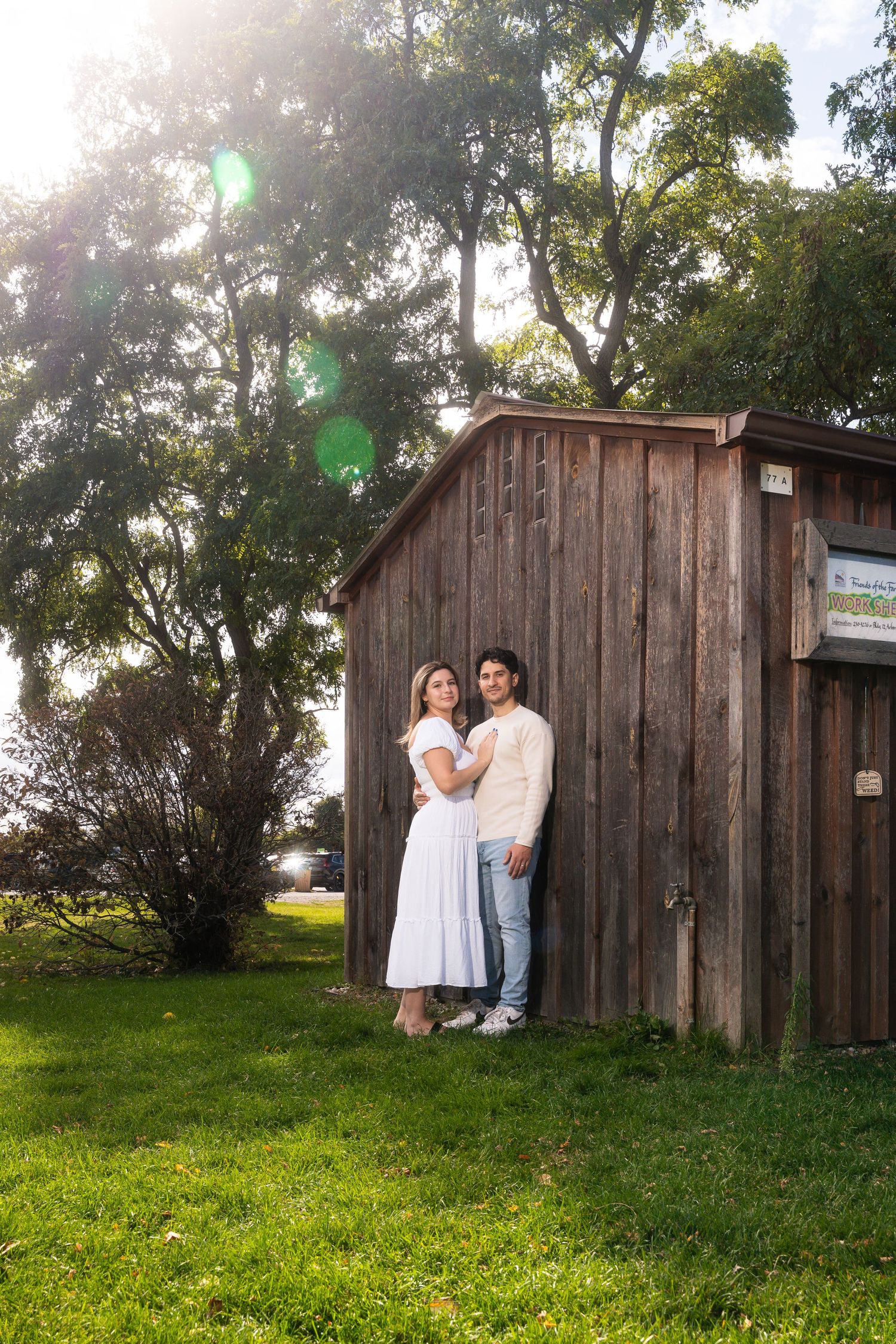 A couple stands together against a rustic wooden barn on a sunny day with green grass and trees in the background.