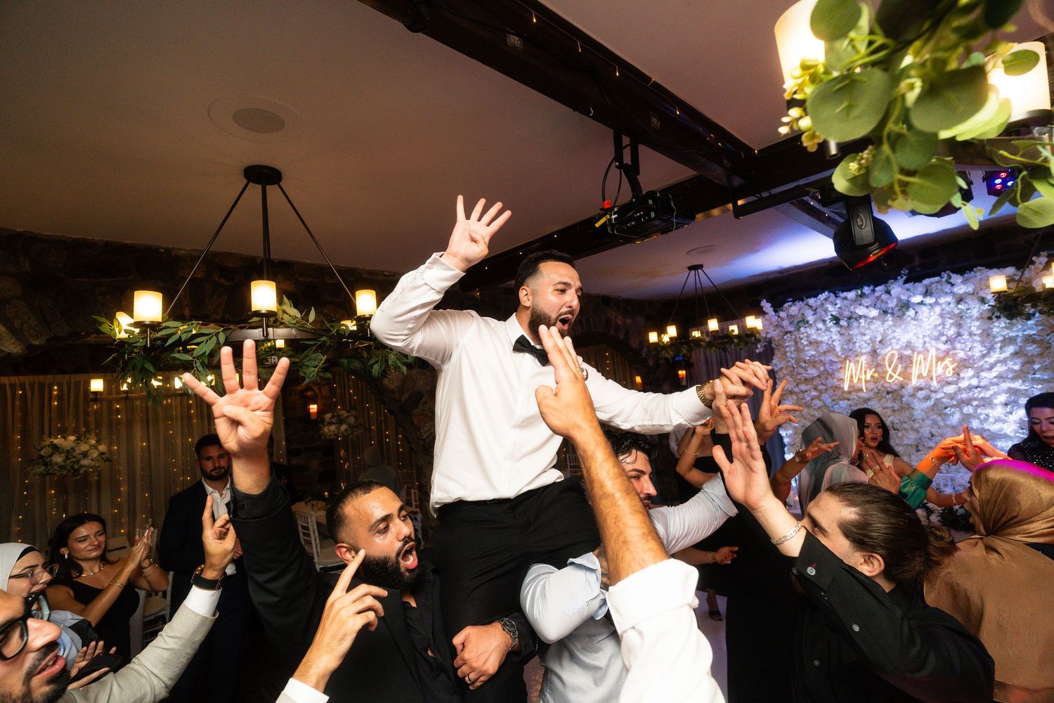 Celebrating guests lift someone in formal attire during an energetic indoor reception party with decorative lighting.