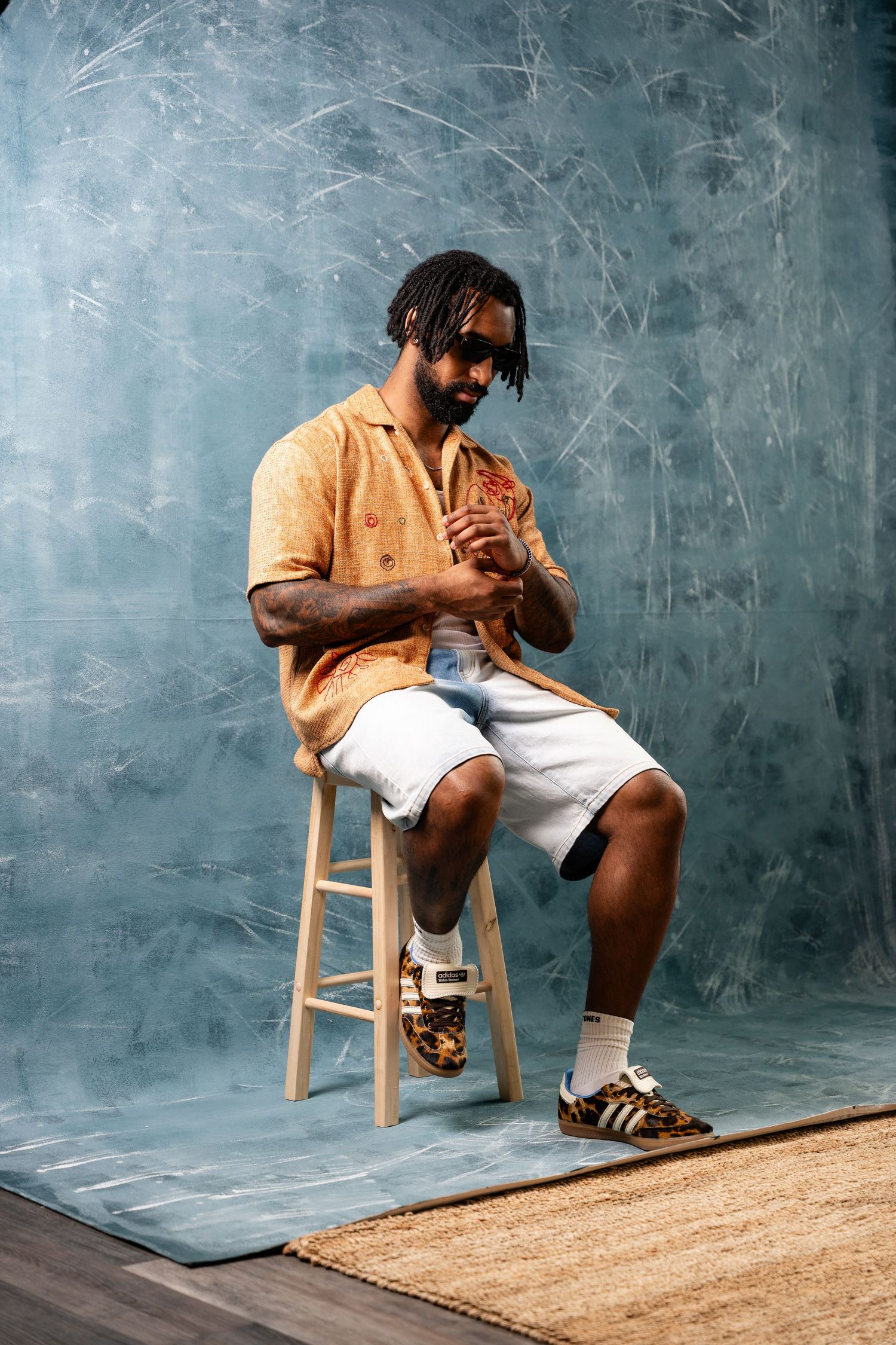Person in yellow shirt and white shorts sits contemplatively on wooden stool against textured grey wall.
