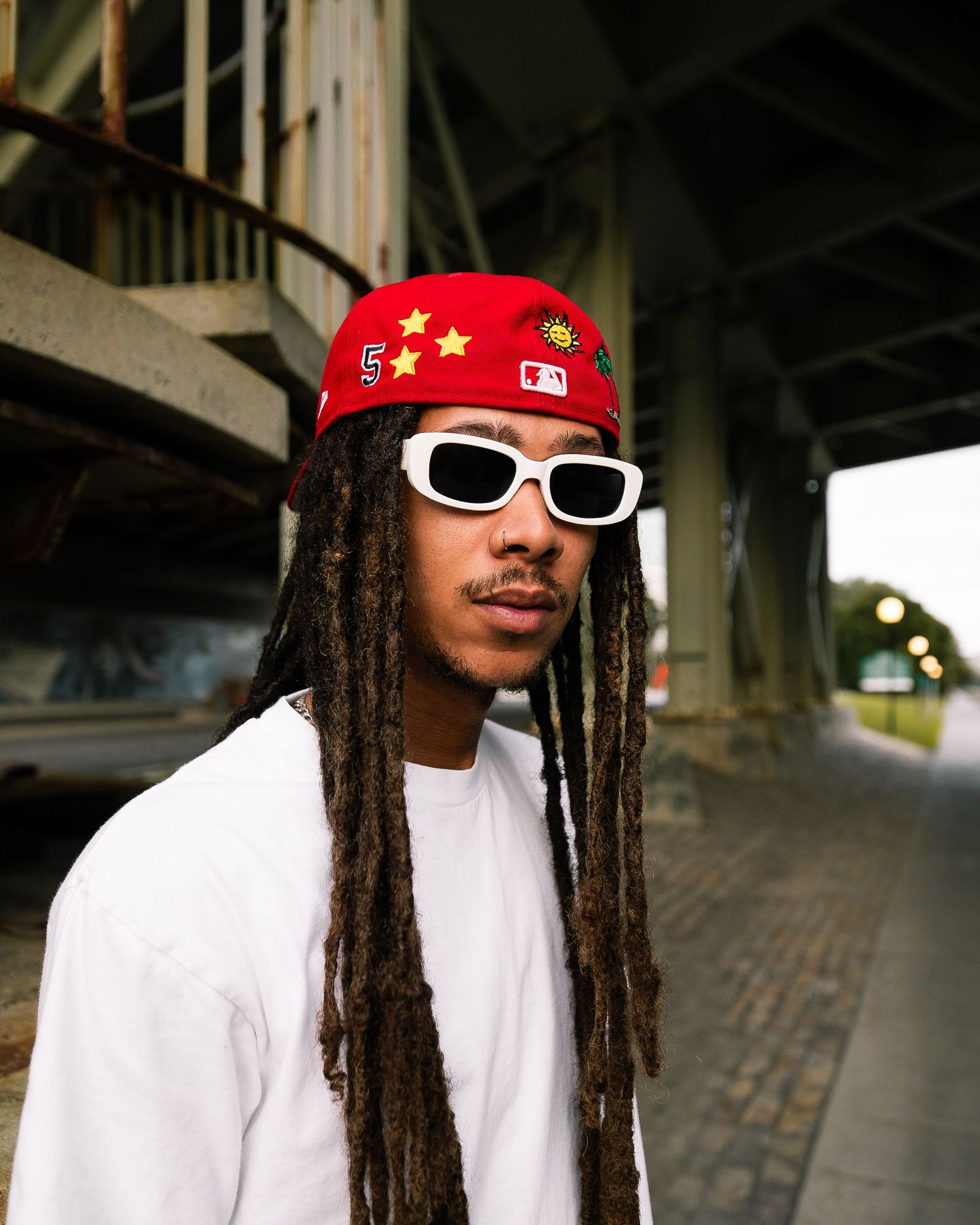 Person wearing white shirt and red cap with white sunglasses under urban bridge.