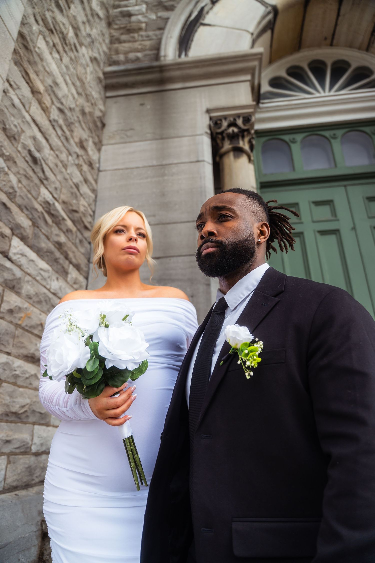 A stylish bride and groom pose outside a historic stone church with ornate green doors and architectural details.