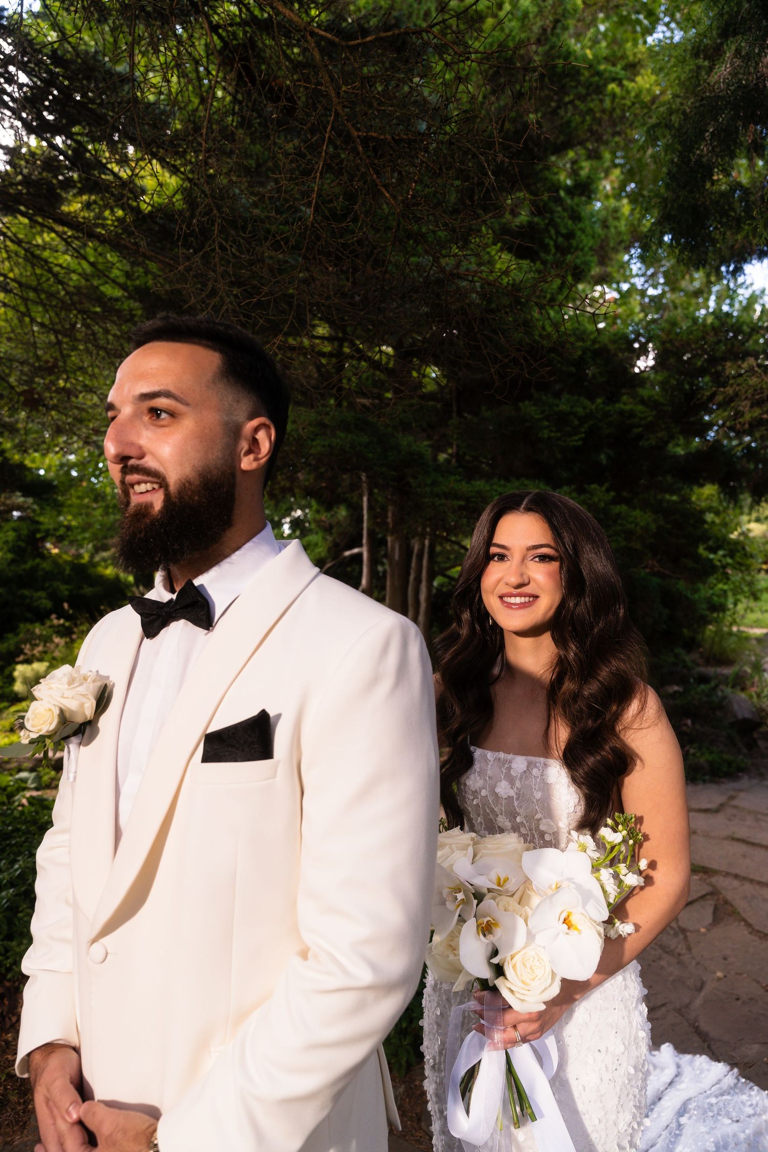 A couple shares romantic moments in their wedding attire with white orchid bouquet in an outdoor garden setting.