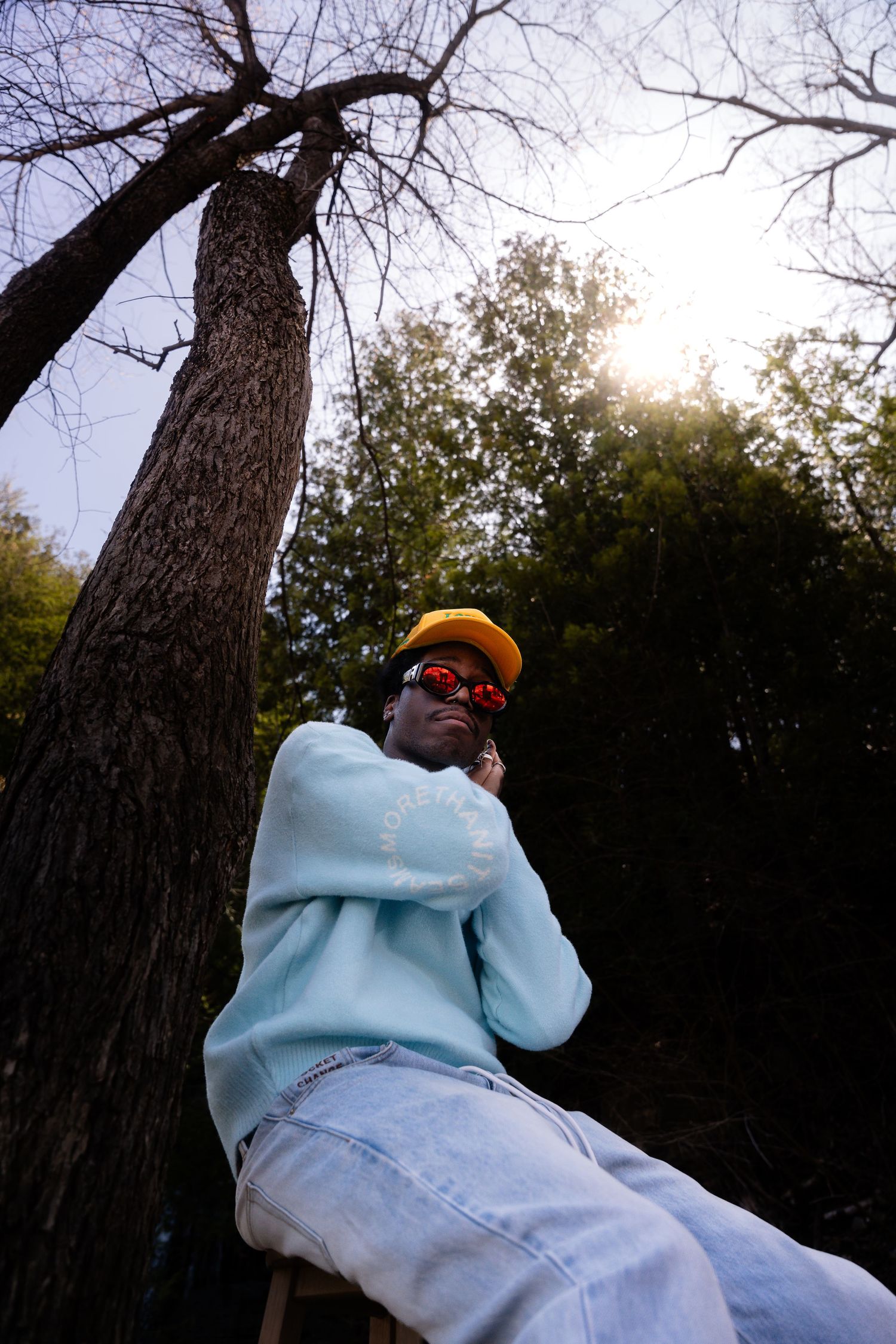 Low angle shot of someone in blue sweater and yellow cap perched by tree.