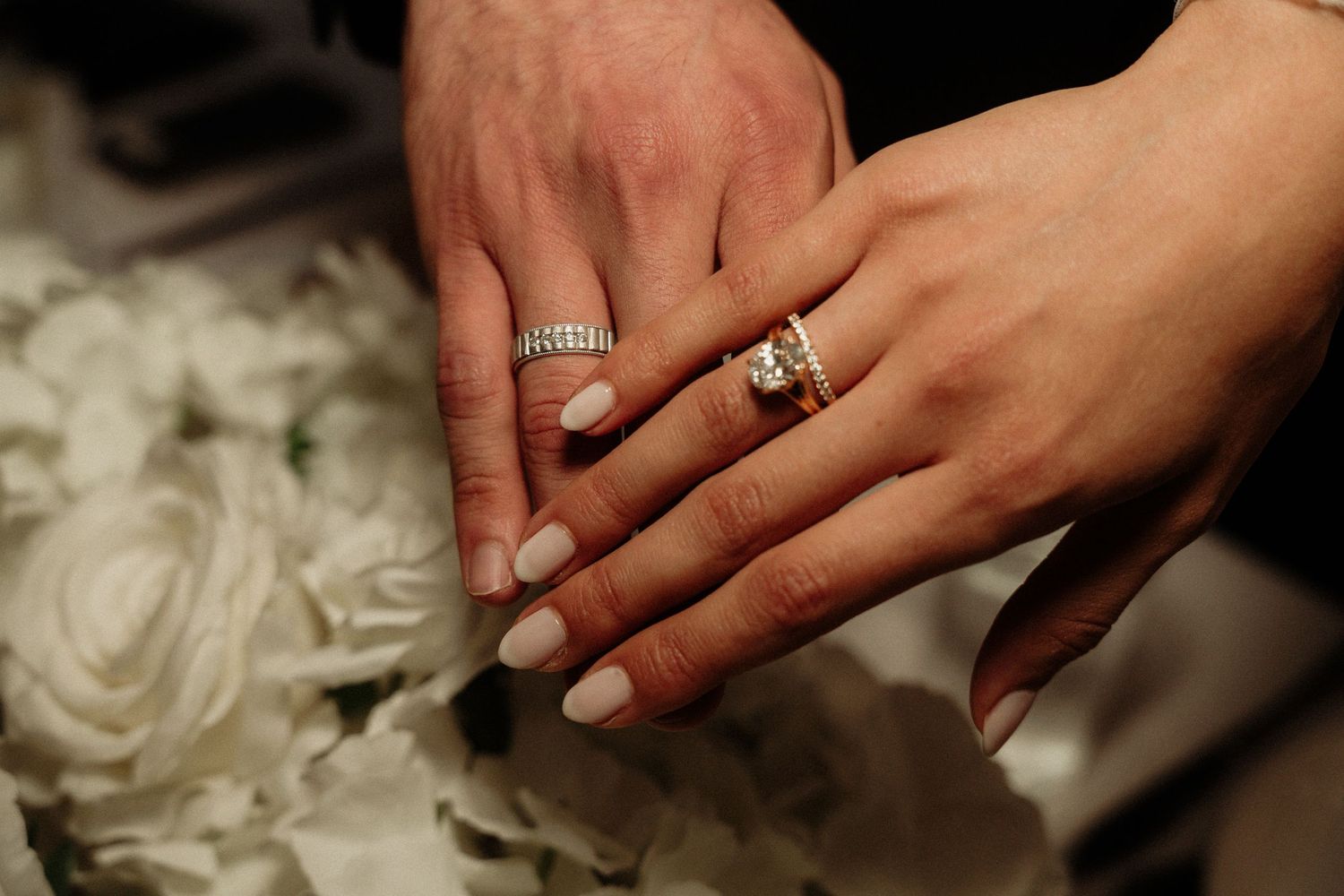 Close-up of wedding bands and engagement rings on hands against textured white material.