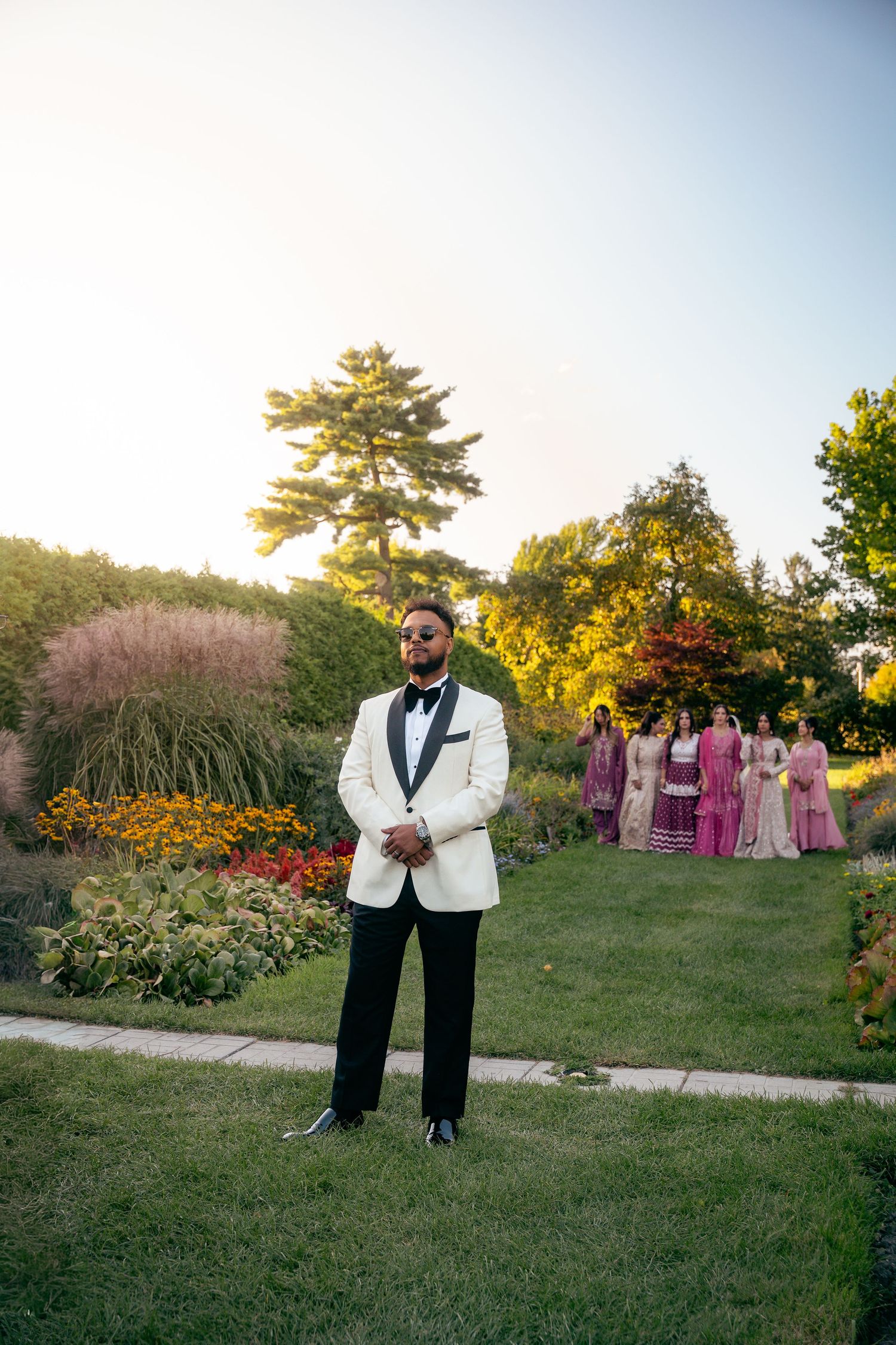 Wedding party poses in garden setting with colorful flowers and greenery during golden hour sunset.