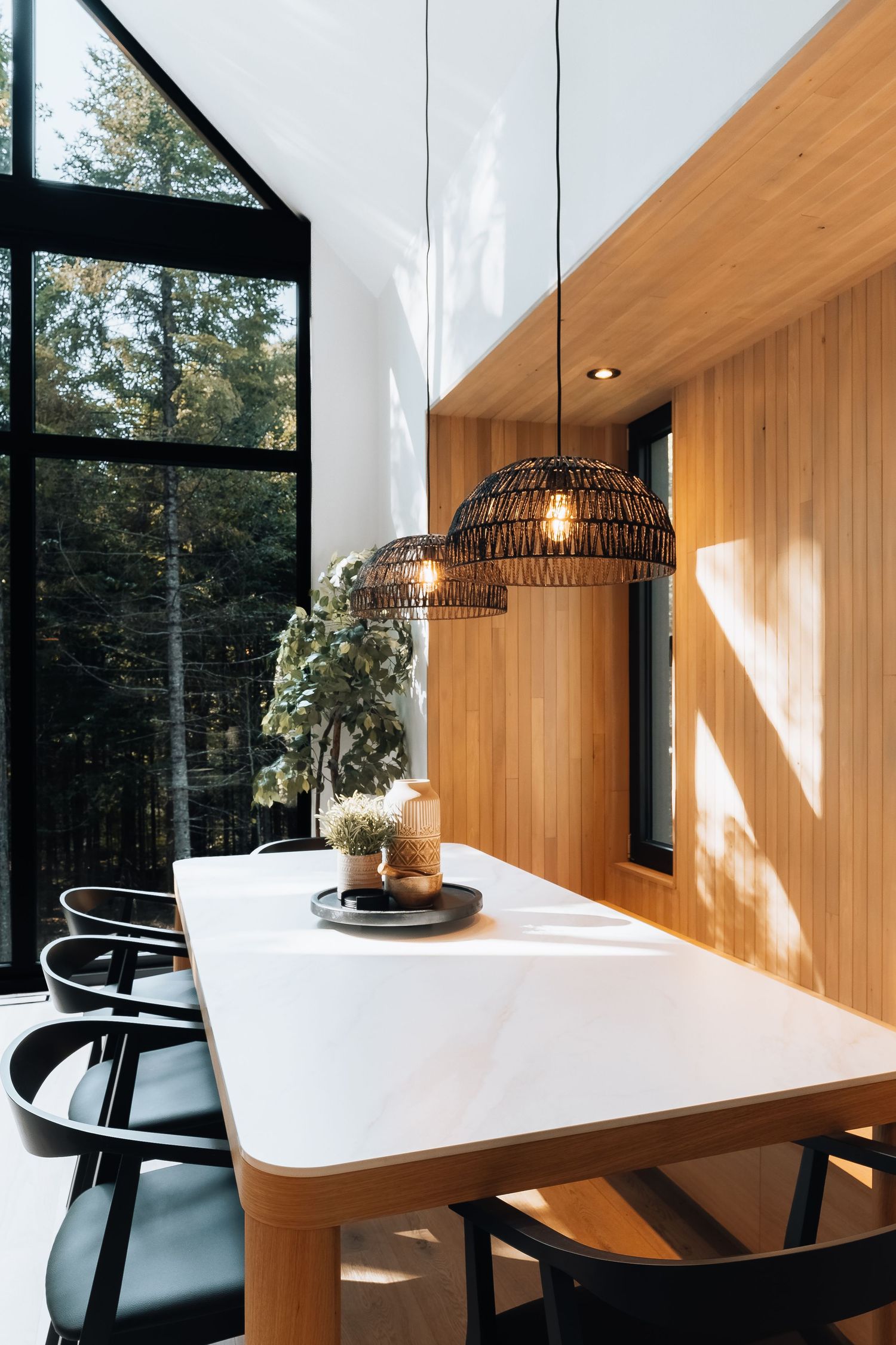 Modern dining space with white table, black chairs, and pendant lights framed by forest views.
