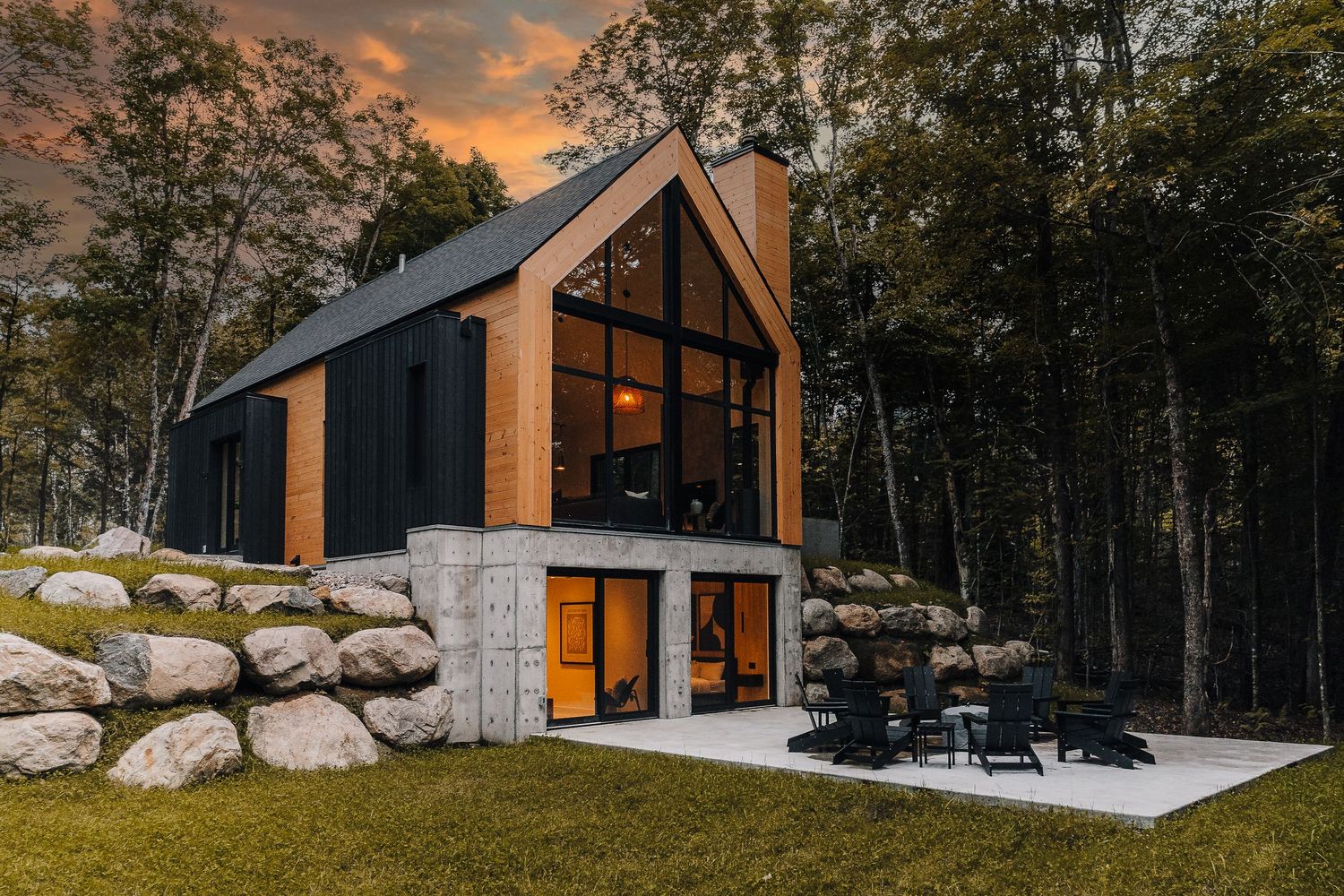 Modern A-frame cabin exterior with large windows and stone accents set against a forest backdrop at sunset.
