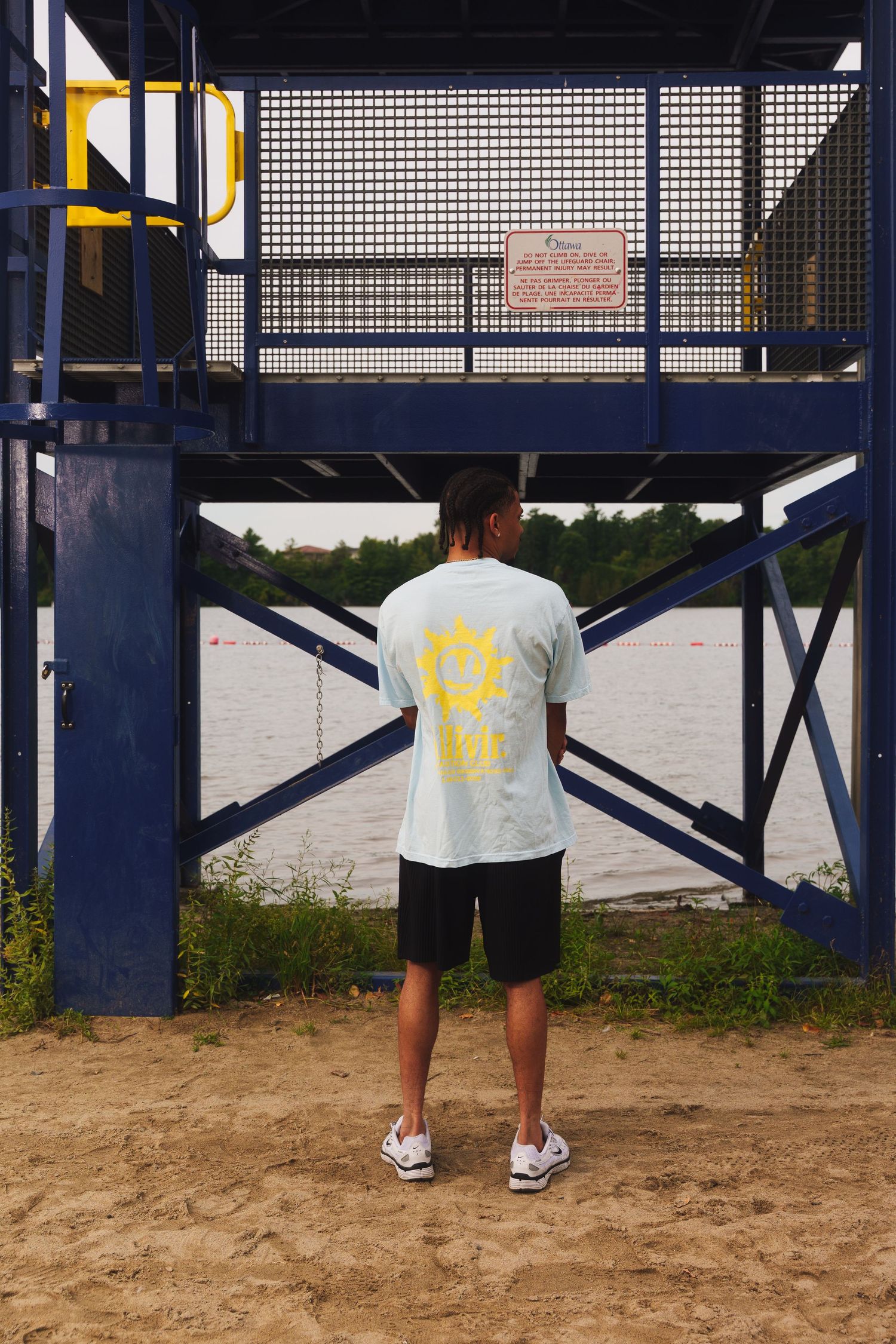 Person in white t-shirt with yellow design standing by blue metal structure near water.