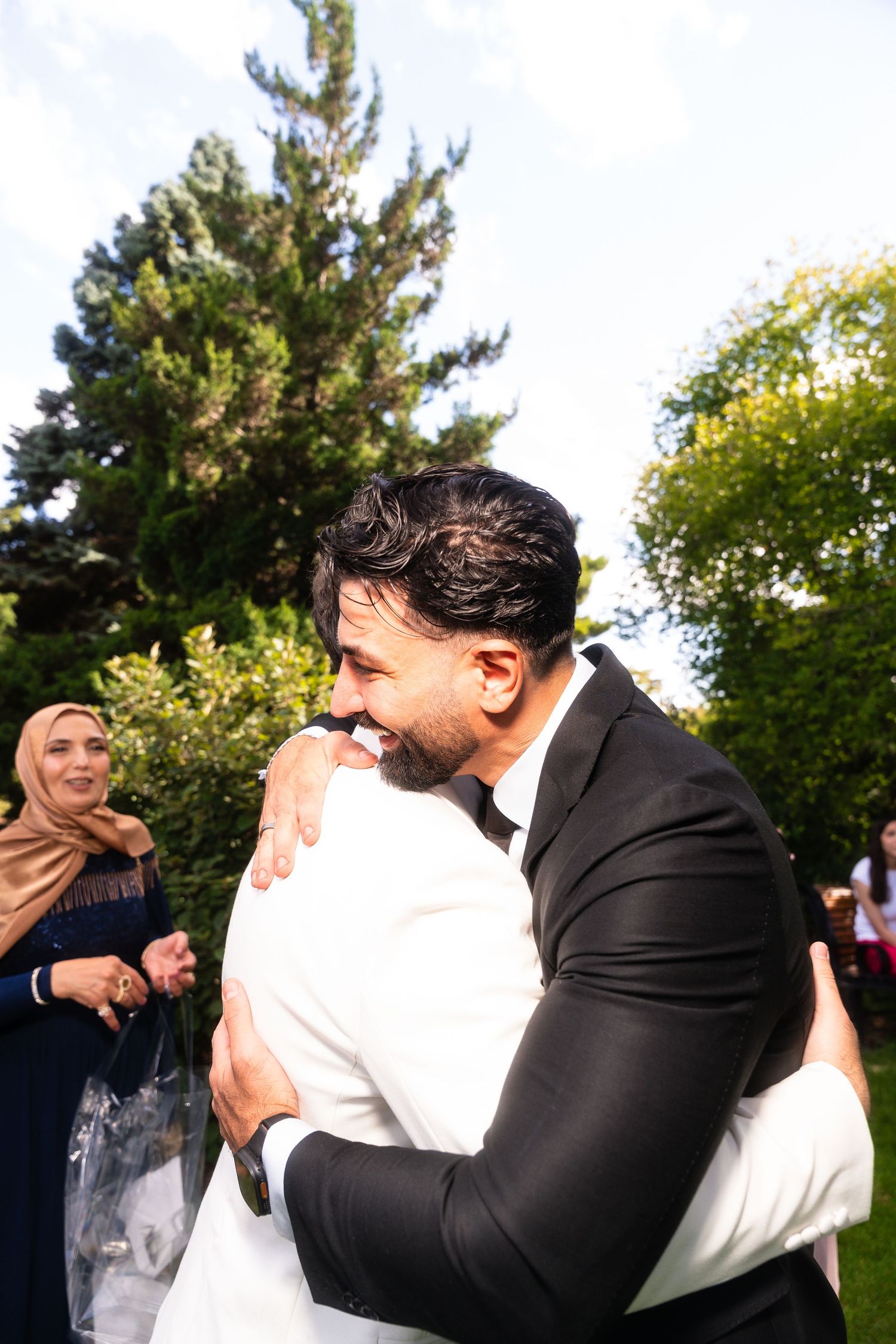 Two people share a warm embrace at an outdoor celebration while guests look on in the background.