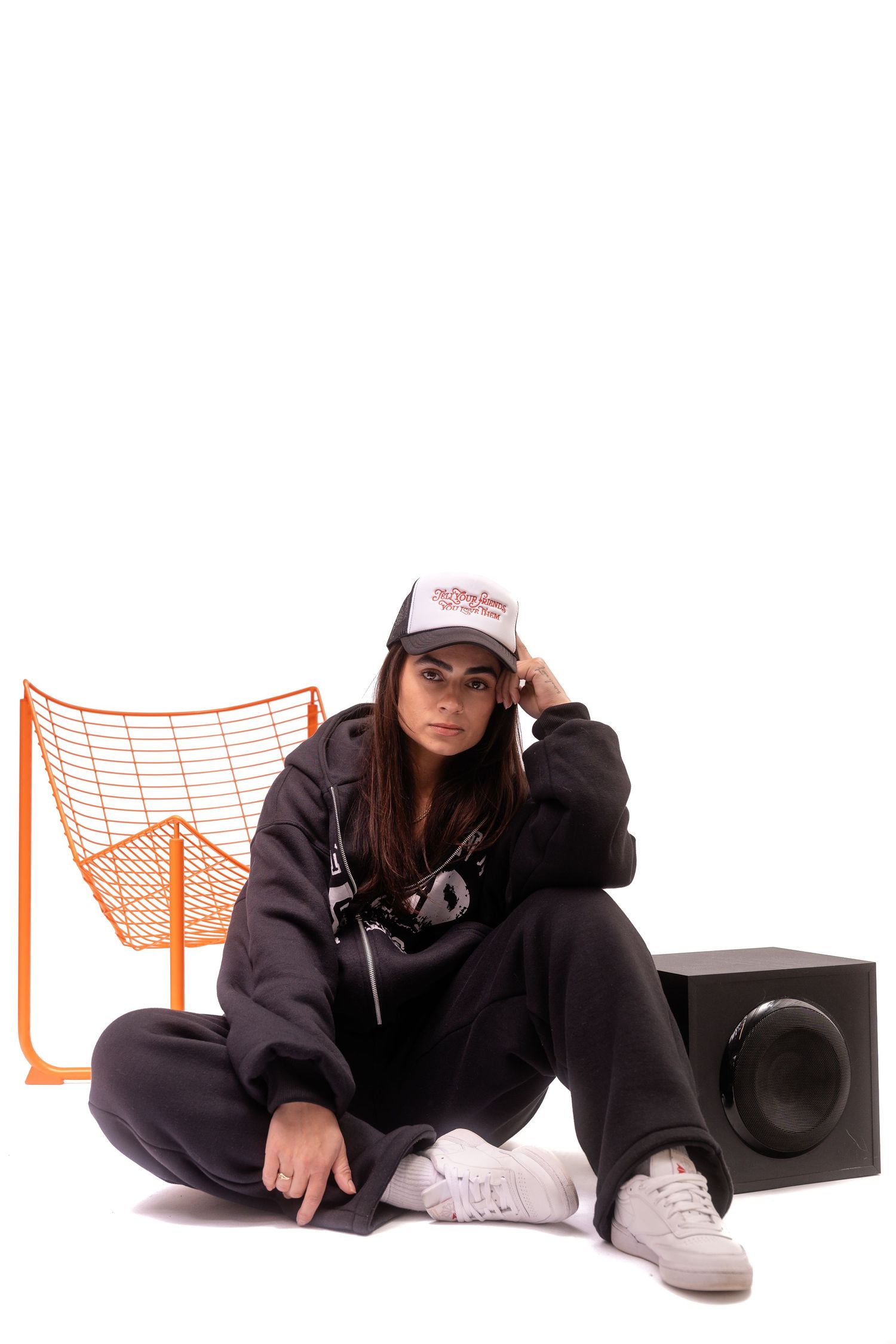 Person in dark hoodie sitting on floor with orange net and speaker in background.